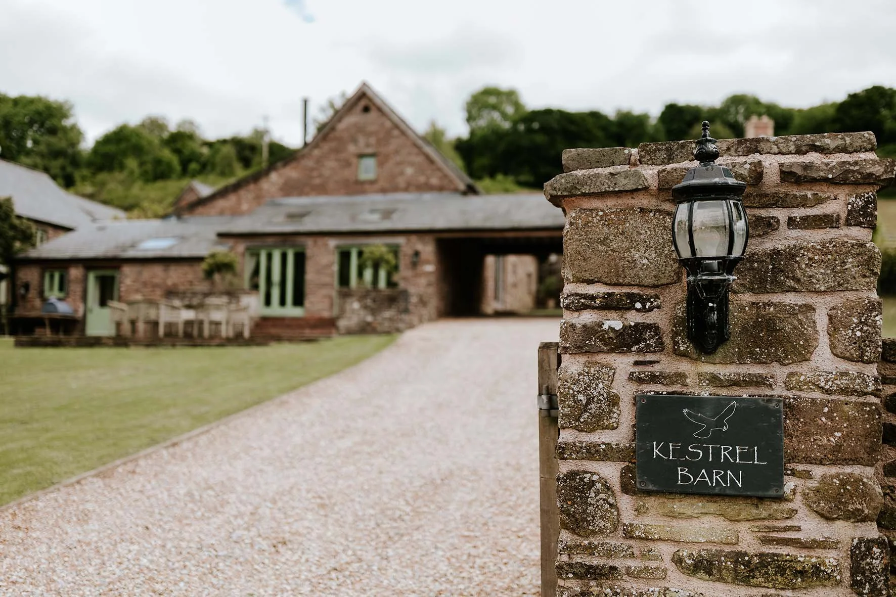 A gravel driveway leading to a stone barn with a pitched roof and multiple windows, surrounded by greenery and trees, with a black lantern attached to a stone pillar and a sign reading 'Kestrel Barn'.
