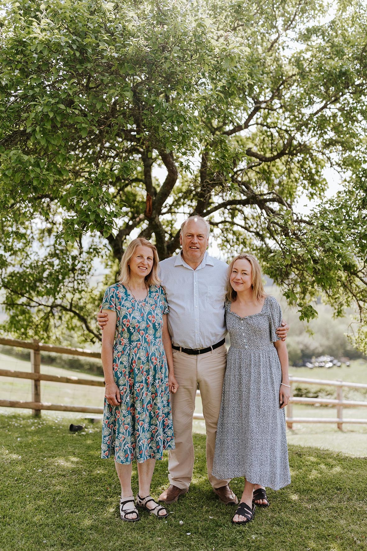 Three people, two women and one man, standing outdoors under a large leafy tree, smiling and embracing each other on a grassy area in a rural setting.