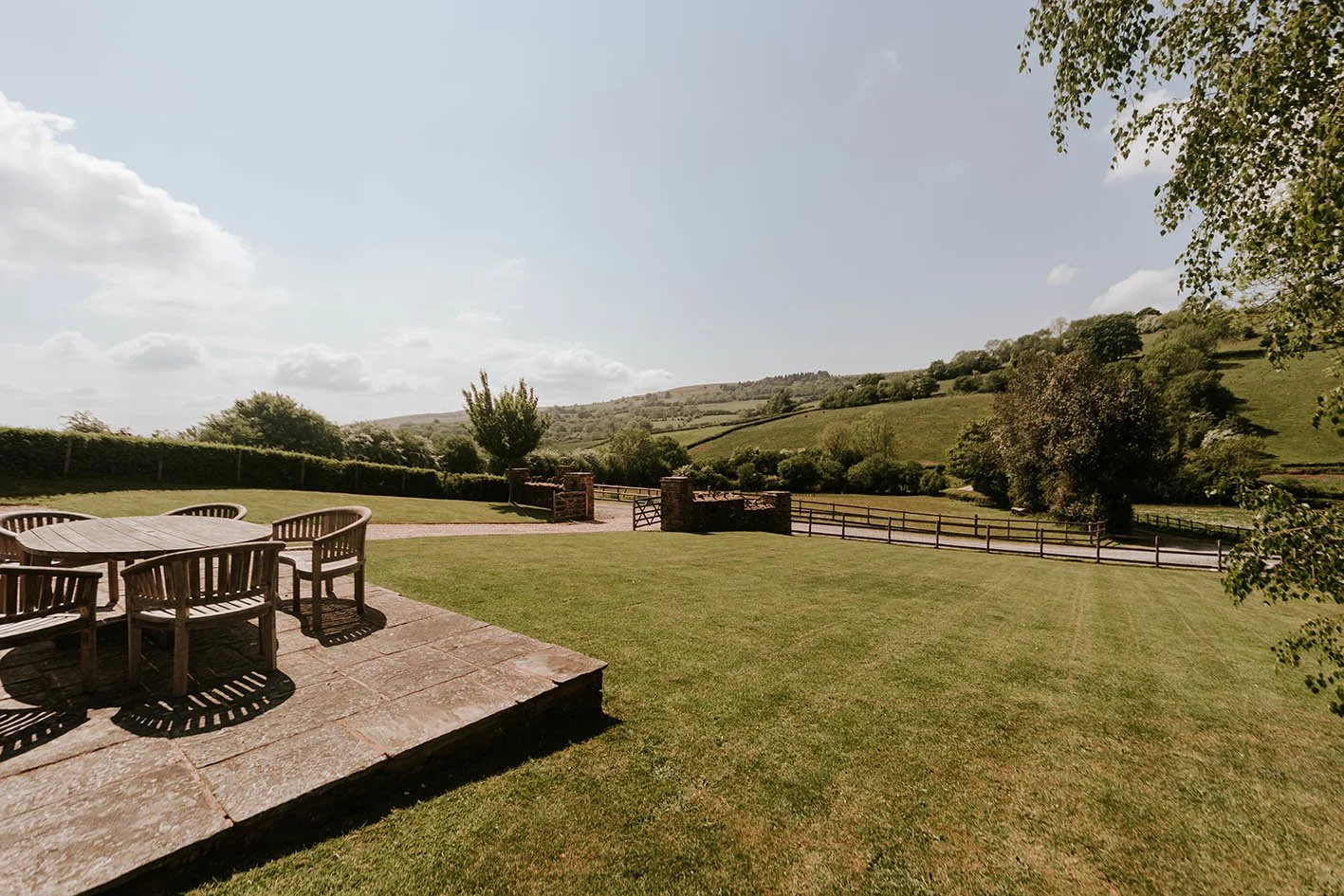 A spacious backyard with a stone patio and wooden outdoor furniture, surrounded by green grass and trees, with rolling hills in the background under a partly cloudy sky.