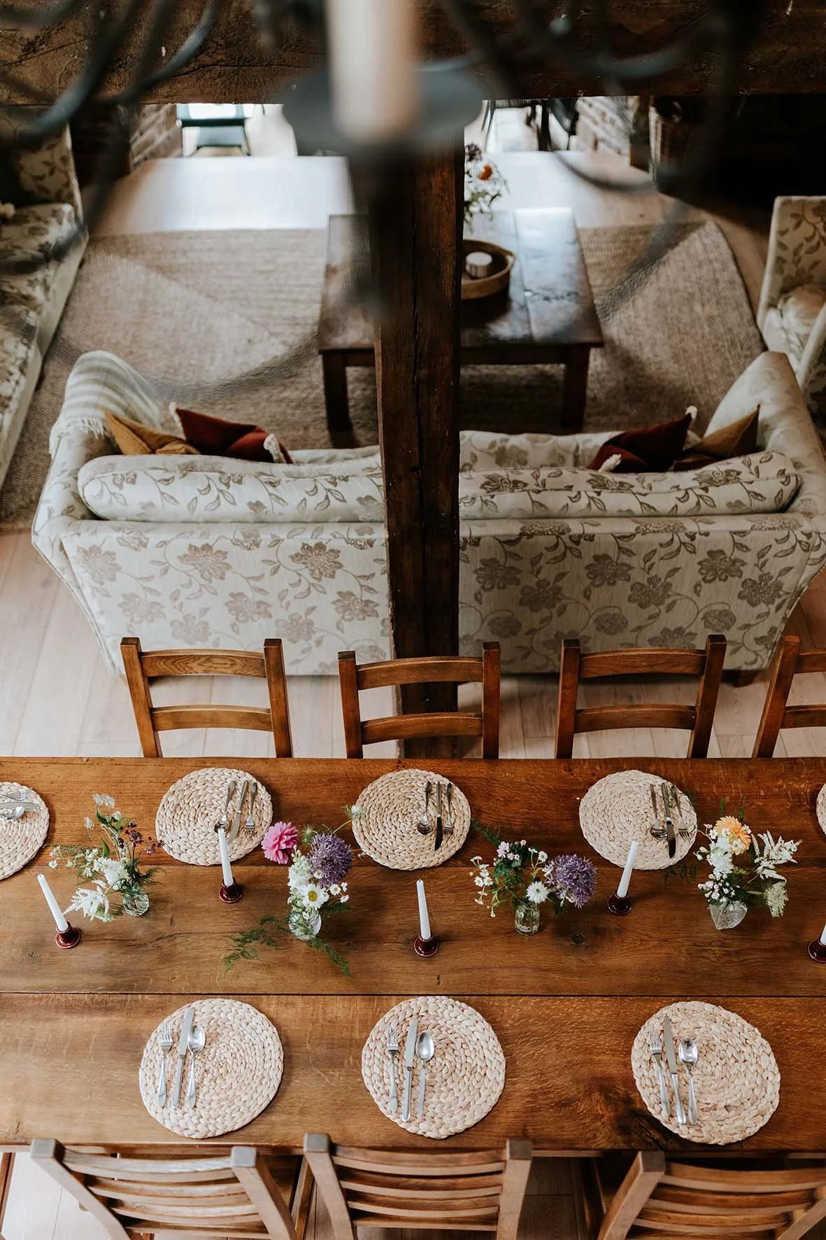 Overhead view of a dining table set with woven placemats, silverware, and small flower arrangements, with a living room and sofa visible through a wooden beam in the background.