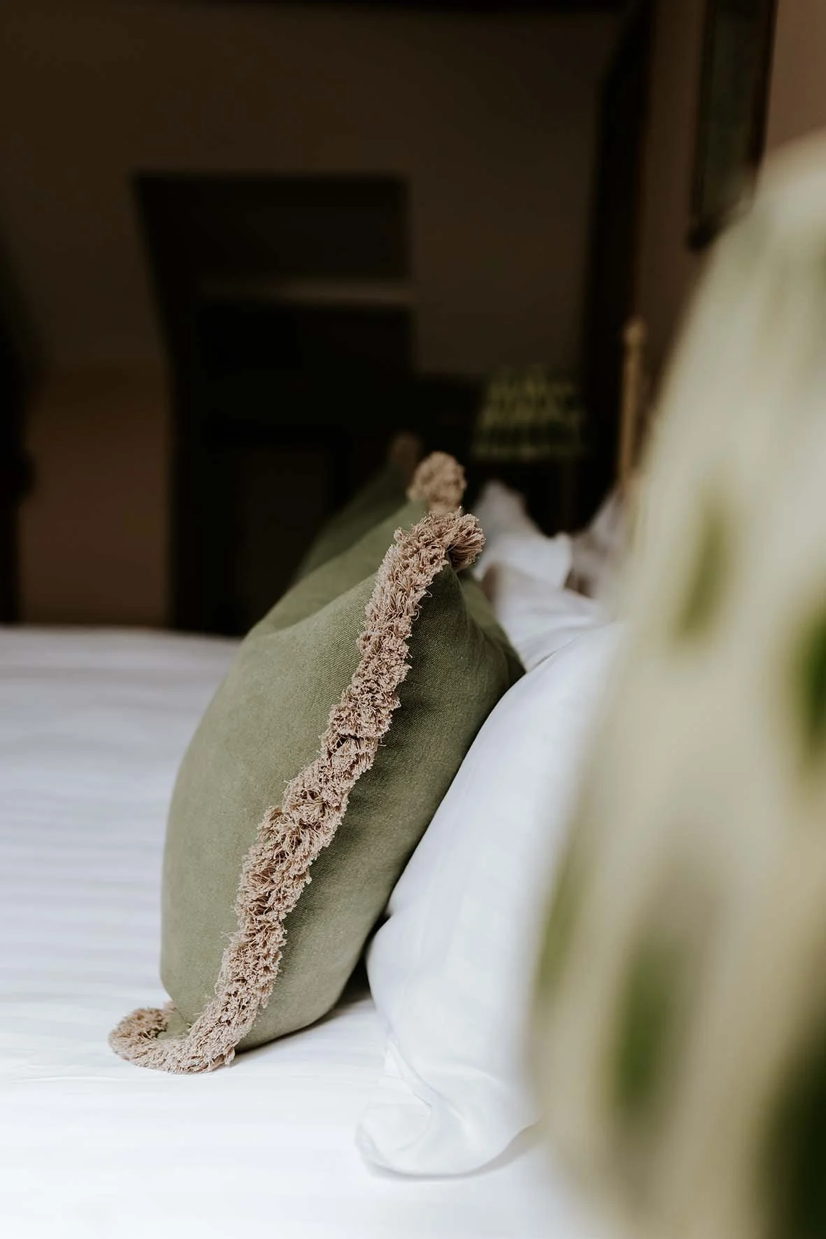 Close-up of a green decorative pillow with beige fringe on a white bed with a white pillow behind it.