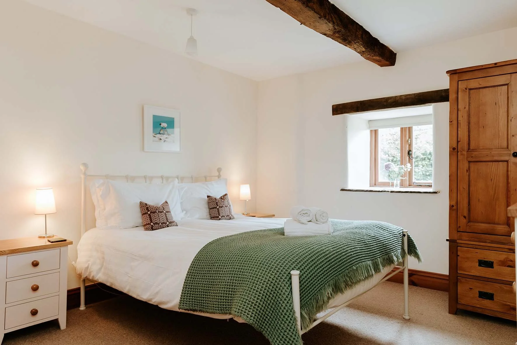 A cozy bedroom with a white bed, green blanket, and rolled towels on the bed. There are two nightstands with lamps, a window with a vase of flowers, and a wooden wardrobe. Exposed wooden beams are on the ceiling.