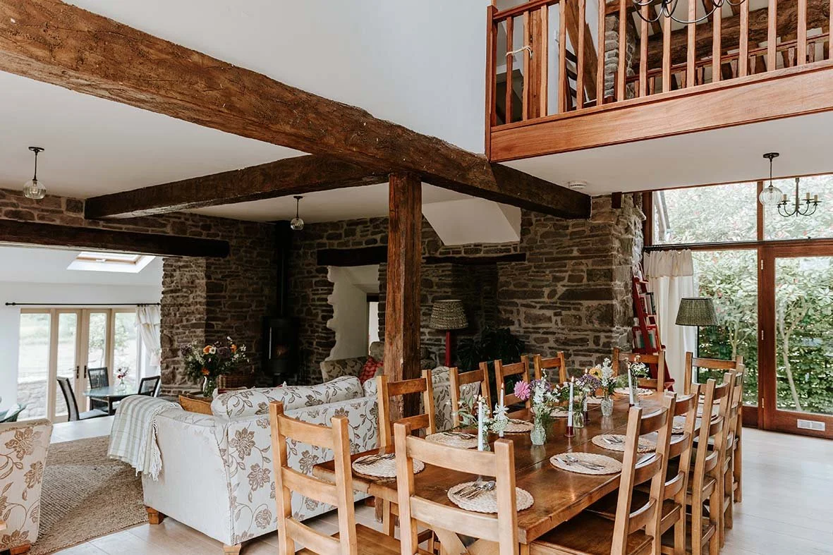 Dining room with a long wooden table, chairs, floral centerpieces, stone walls, large windows, and exposed wooden beams.