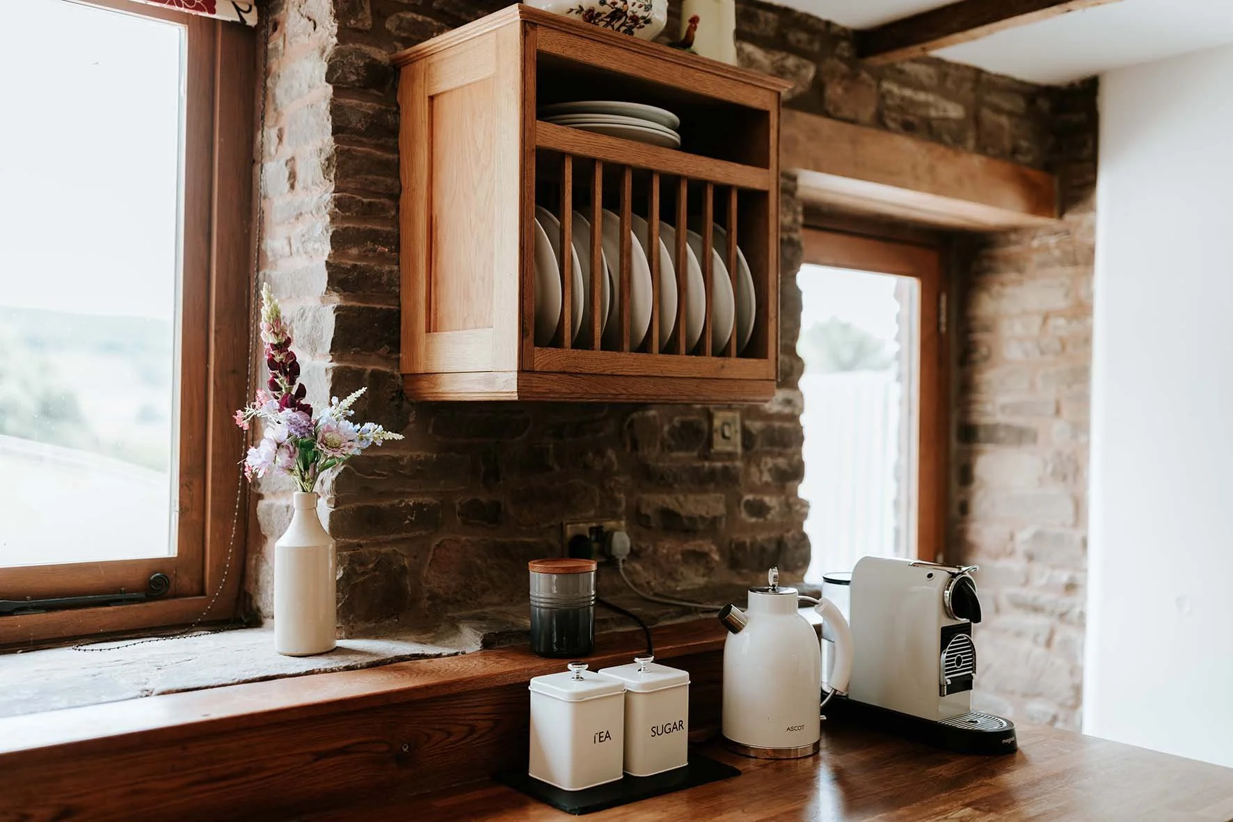Kitchen counter with white ceramic containers labeled tea and sugar, a white electric kettle, and a coffee machine. A small vase with pink artificial flowers is near the window. A wooden wall cabinet holds neatly stacked plates. The background featur