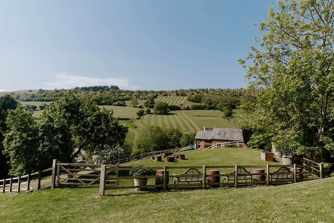 Scenic view of a farm with green rolling hills, trees, a wooden house, and a fence in the countryside under a clear blue sky.