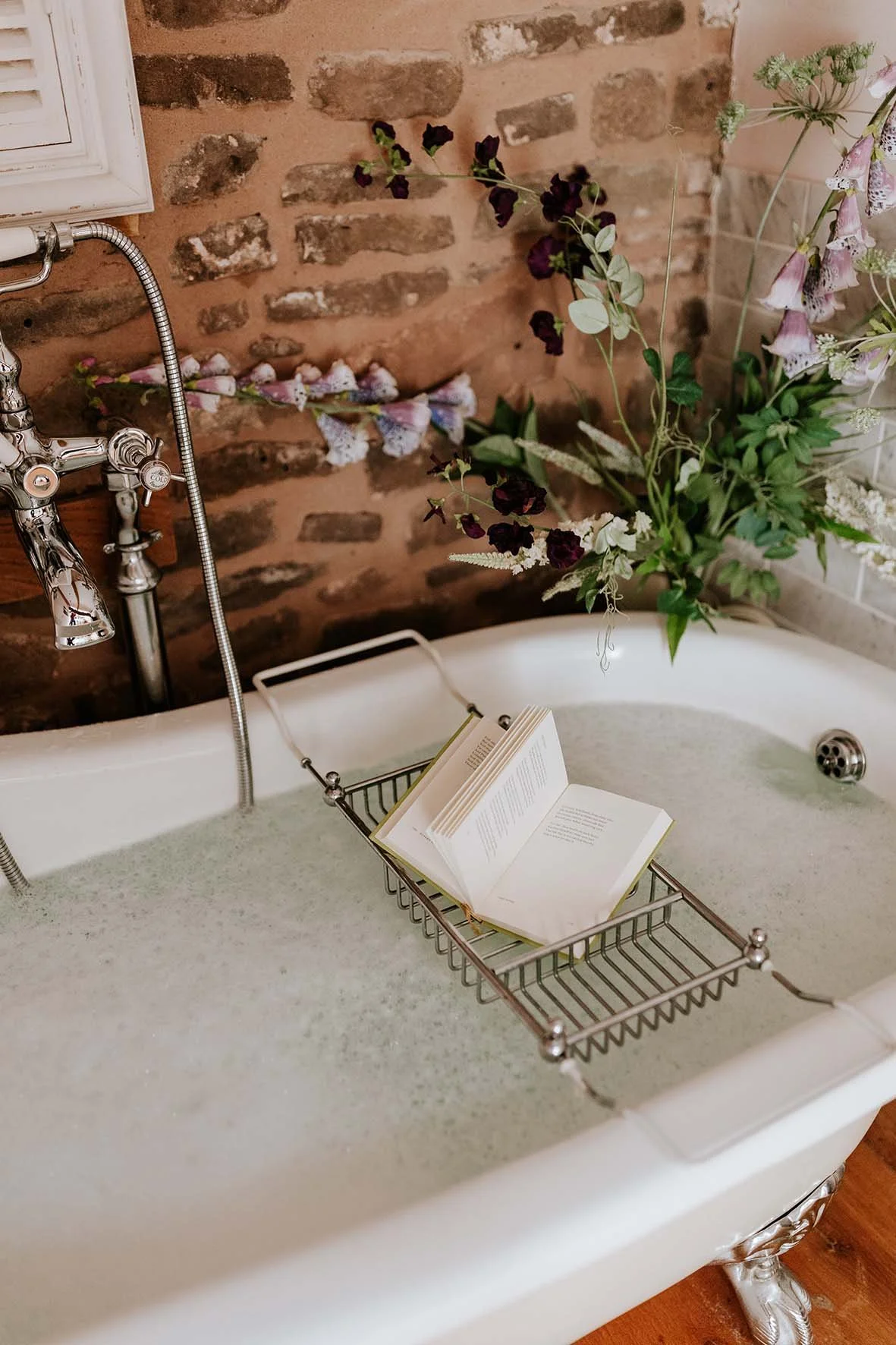 An antique clawfoot bathtub filled with water, with an open book resting on a metal bath rack inside it, and a brick wall with hanging flowers in the background.