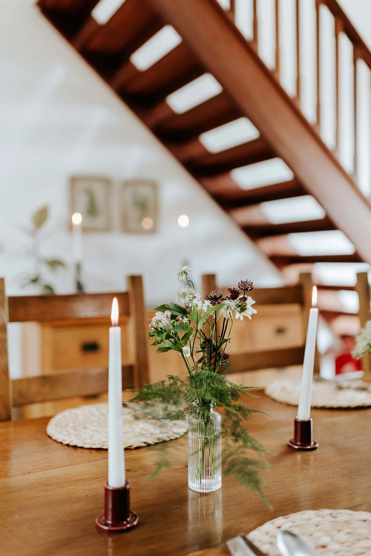 A wooden dining table with a floral centerpiece in a glass vase, surrounded by candles and woven placemats, inside a house with a staircase in the background.