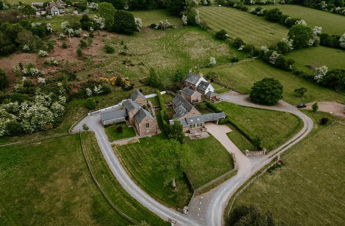 Aerial view of a large stone house with multiple wings, surrounded by a gravel driveway, green lawns, trees, and fields.