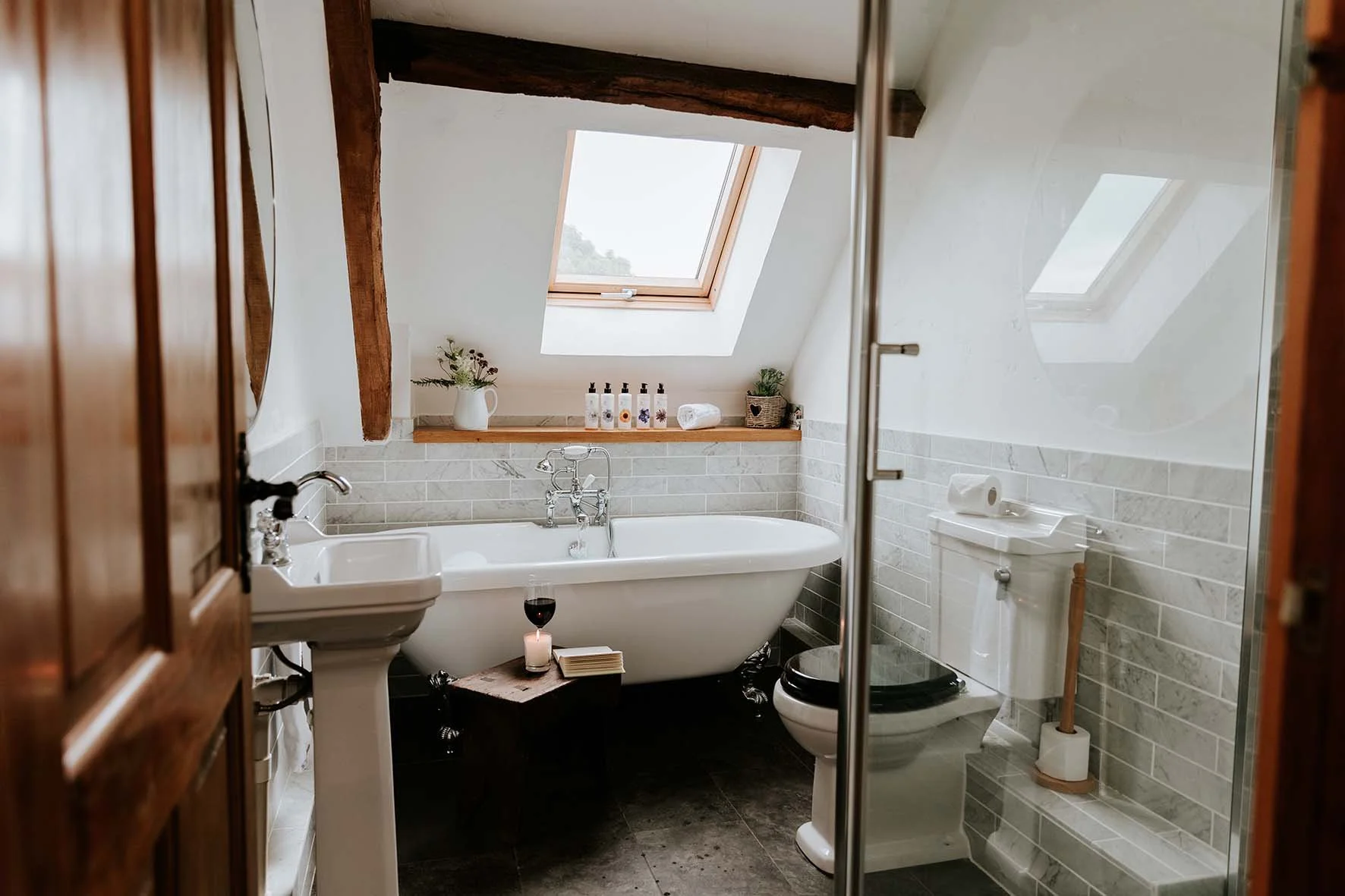 Bathroom with a bathtub under a skylight window, a white pedestal sink, a toilet, a wooden door, and natural light coming through the skylights.