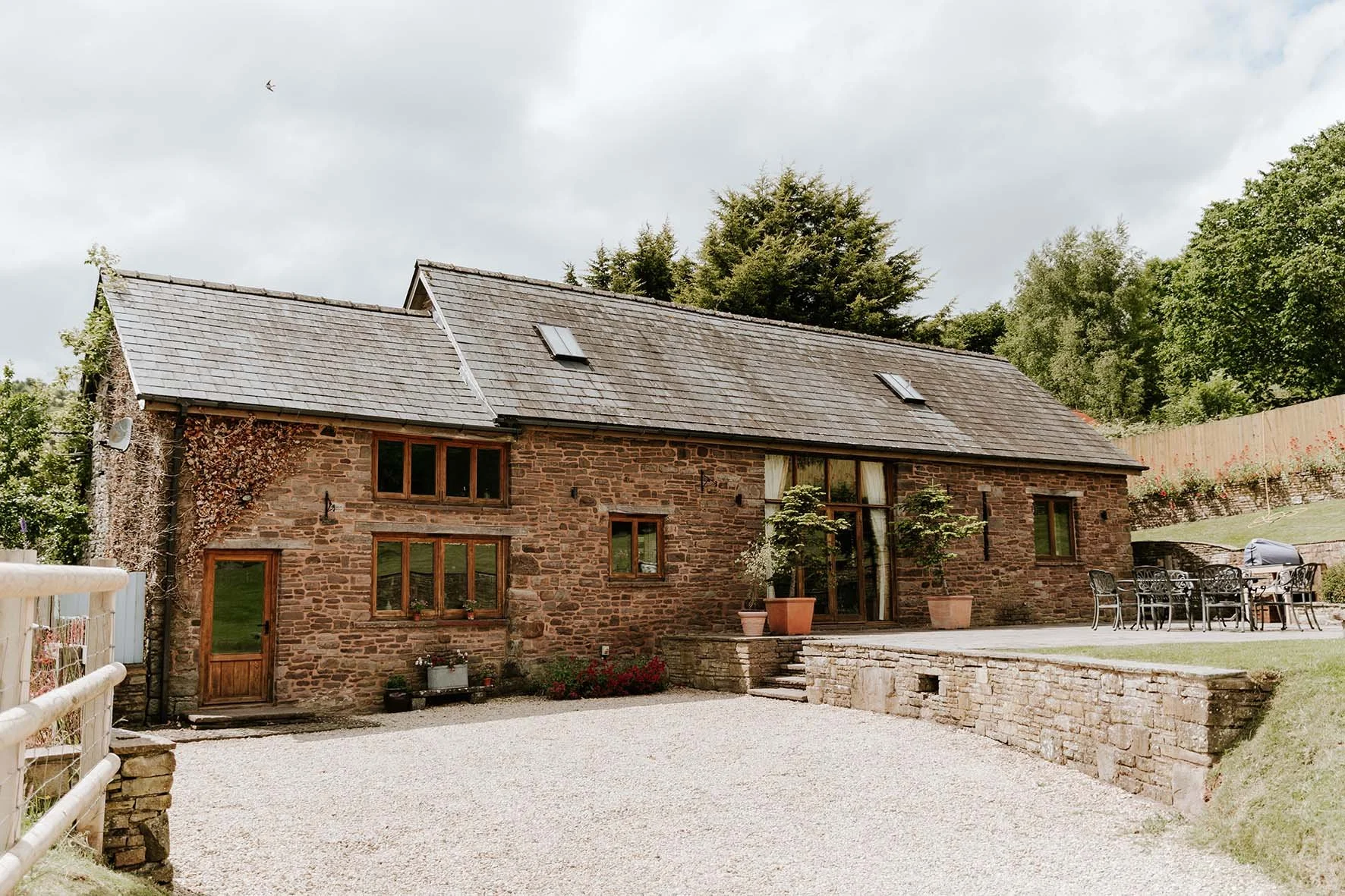 Stone house with a sloped roof, multiple windows, an outdoor dining area, and potted plants in a garden setting.