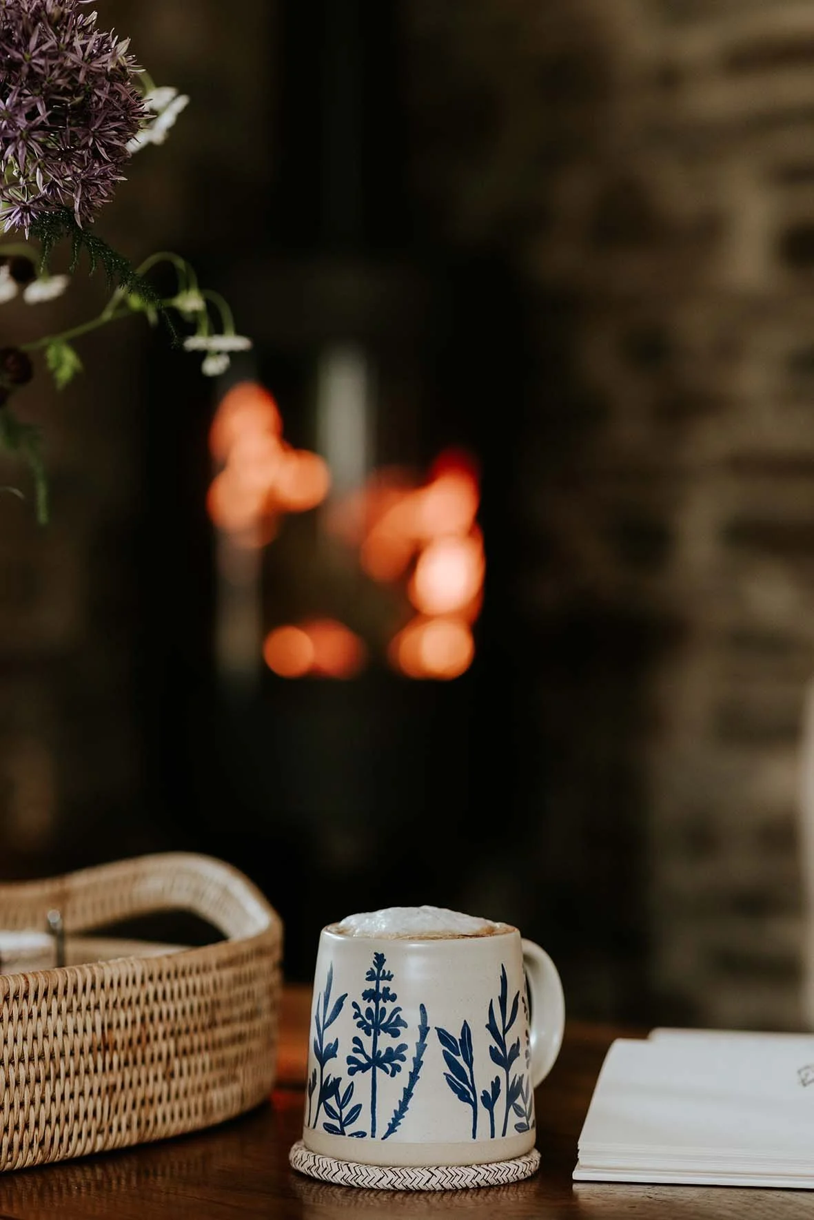 A mug with a blue floral pattern on a table next to a wicker tray and some notebooks, with a fireplace burning in the background.