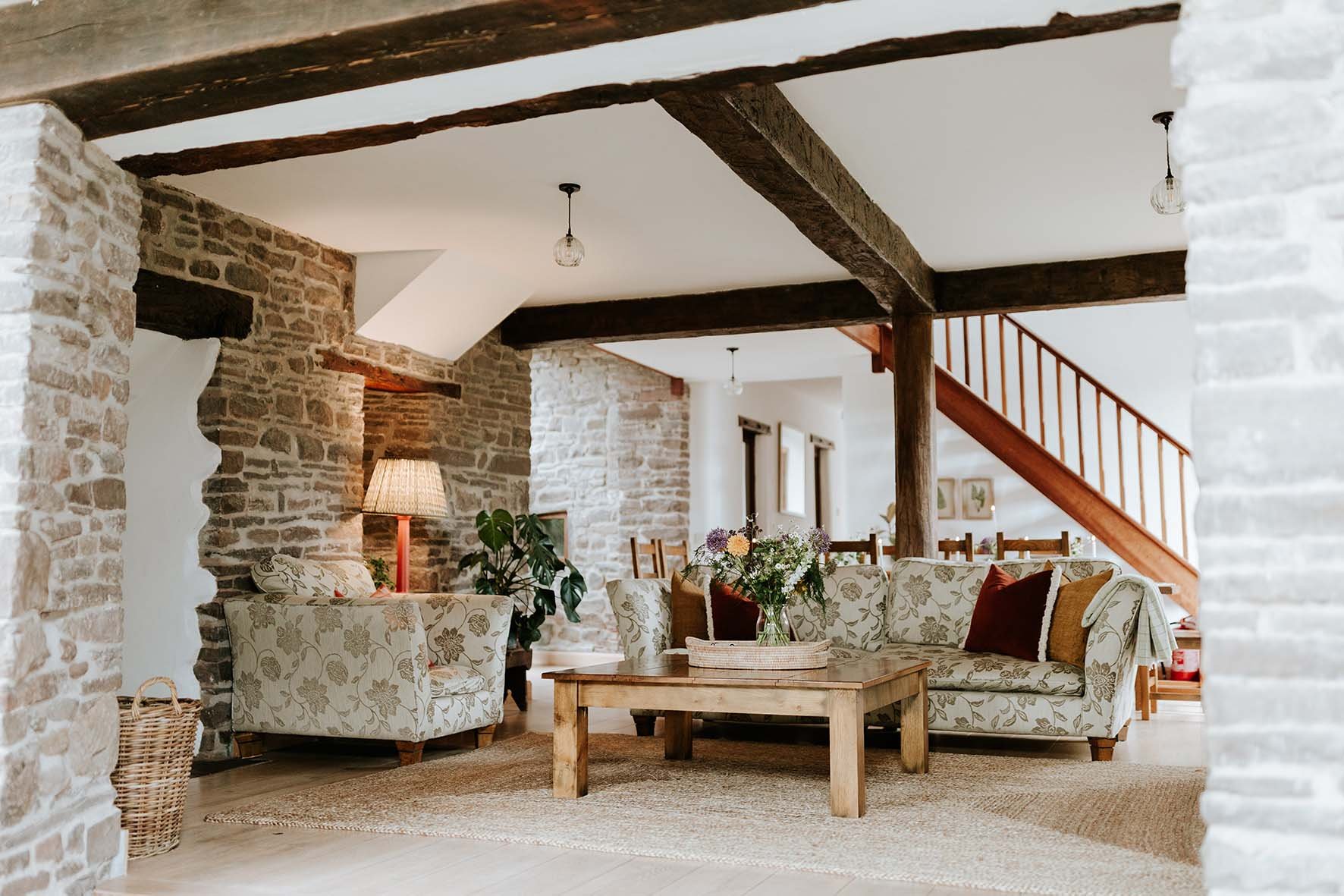 Living room with stone walls, dark wooden beams, floral sofa, armchair, wooden coffee table with flower arrangement, floor lamp, and staircase in the background.