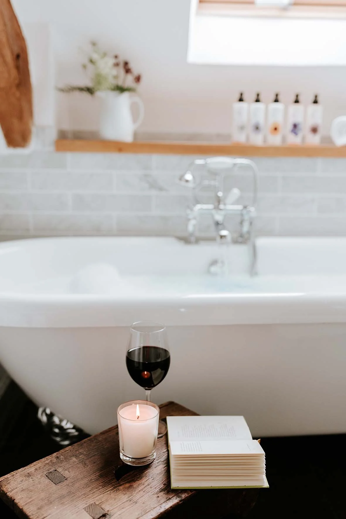 A wooden table with a candle, a glass of red wine, and an open book in front of a bathtub with a faucet, in a bathroom setting with a shelf holding a white pitcher with flowers and bottles, and a window in the background.
