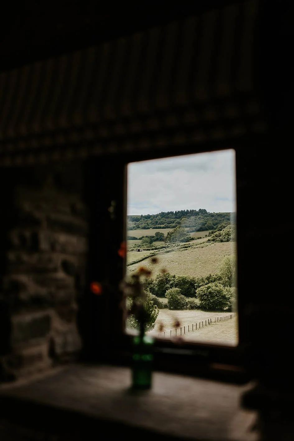 View of green, rolling countryside through a small window, with fields, trees, and a distant hill under a partly cloudy sky.