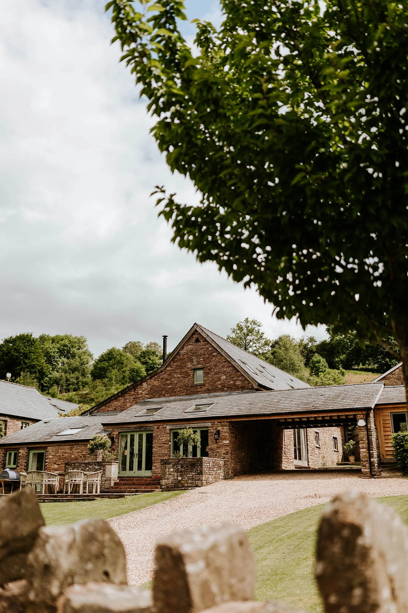 A rustic house made of stone with a slate roof, situated on a green lawn with a gravel driveway in front, surrounded by trees under an overcast sky.