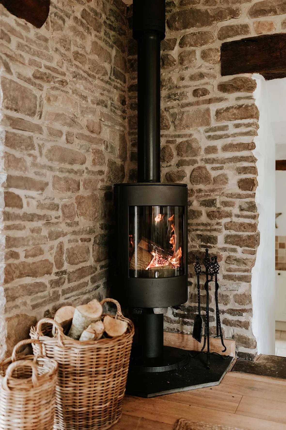 A wood-burning stove with a glass door showing flames inside, situated against a rustic brick wall. To the left, there is a basket filled with firewood, and to the right, a set of fireplace tools.