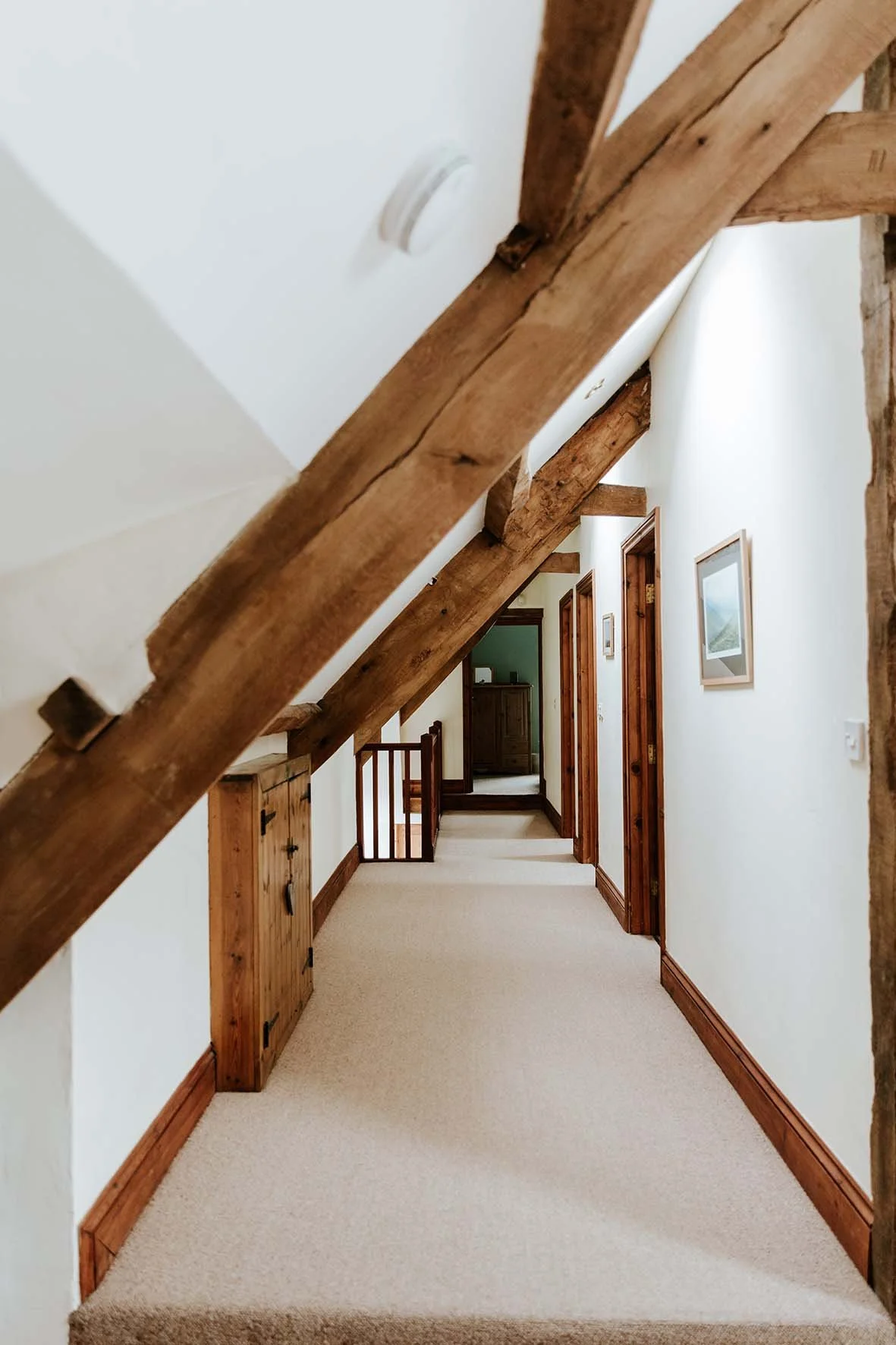 Hallway with white walls, wooden trim and beams, carpeted floor, framed picture on wall, and doors leading to other rooms.
