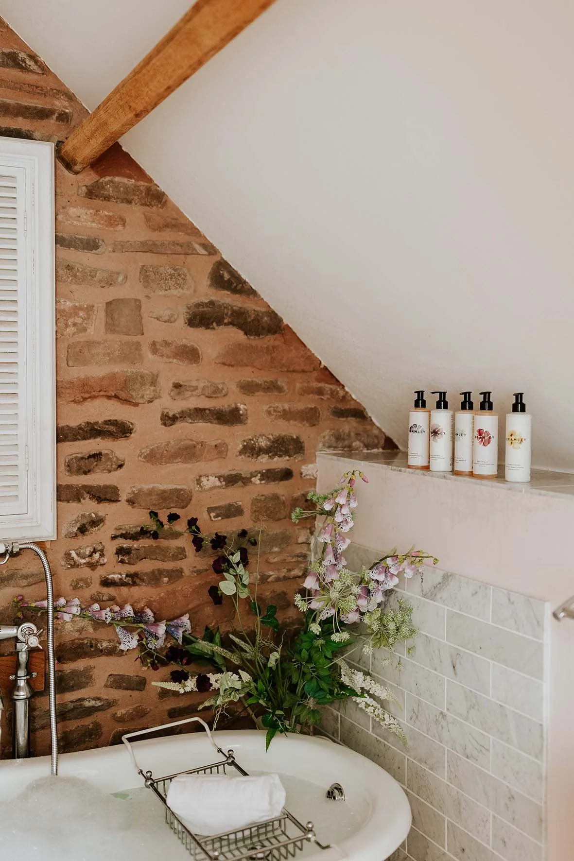 Decorative bathroom scene with a bathtub, a flower arrangement, and skincare bottles on a ledge. Exposed brick wall and sloped ceiling.
