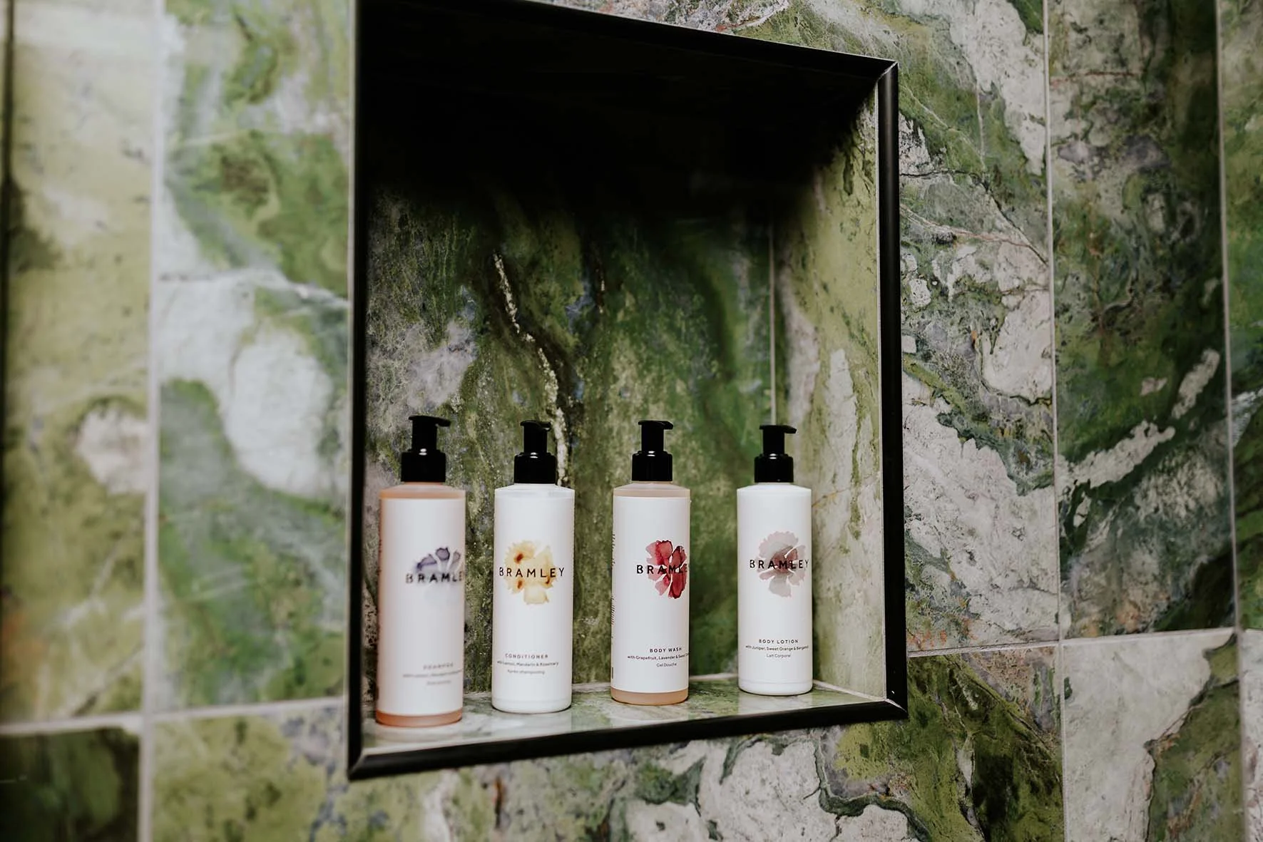 Four bottles of Bramley lotion and body wash on a green marble tiled shelf in a shower or bathroom wall niche.