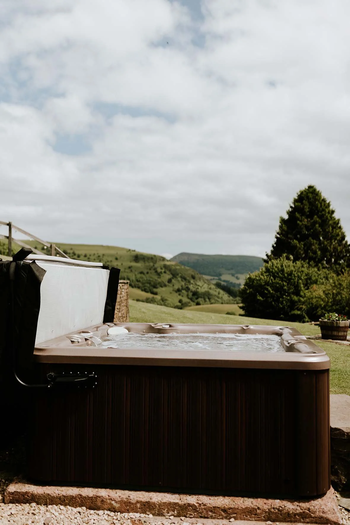 Outdoor hot tub on a grassy hill with scenic rolling hills, a large tree, and cloudy sky in the background.