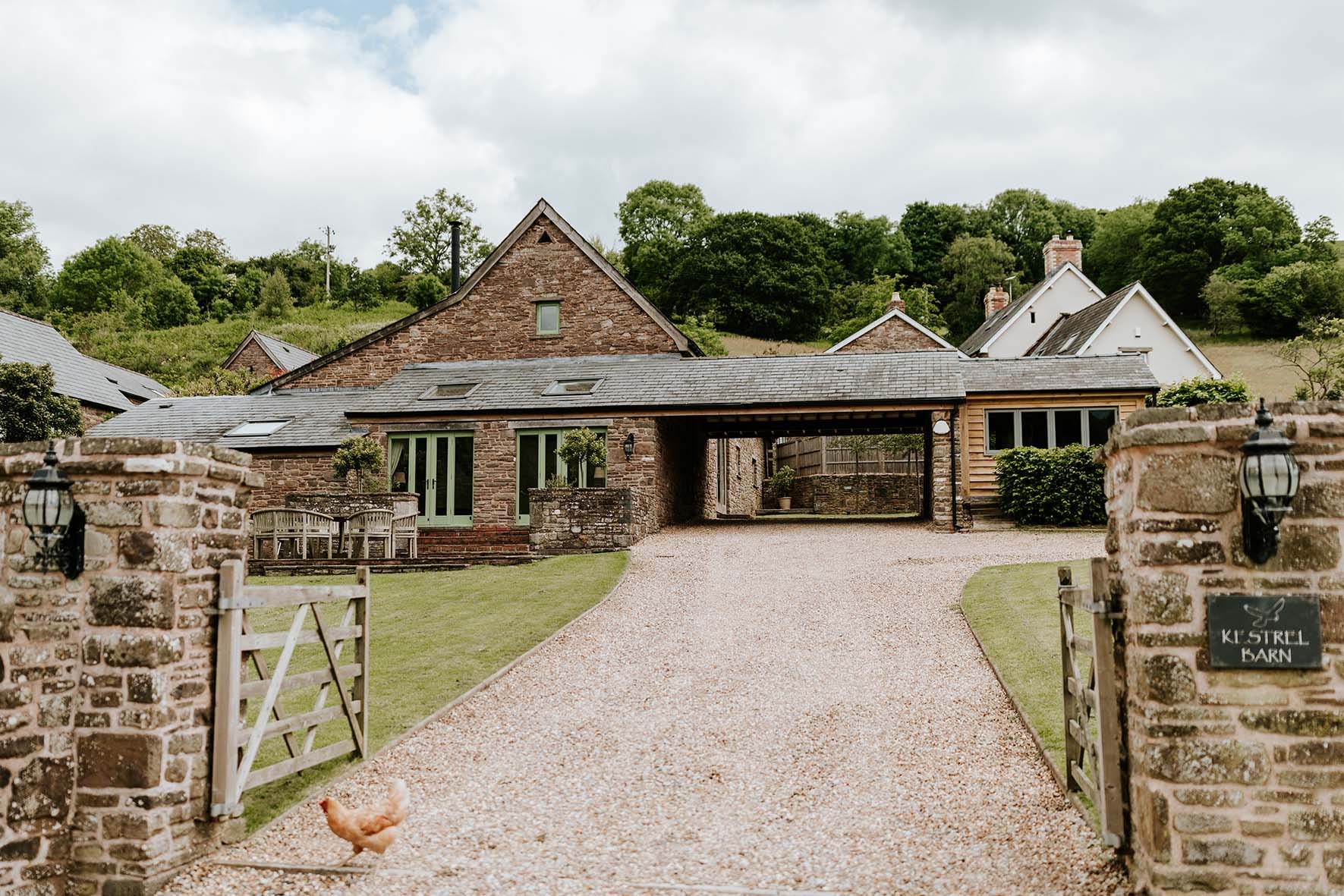 A rustic stone house with a covered driveway, surrounded by a gravel path, green lawn, and stone fences. A hen is near the gate, and a sign reads 'Ki Strel Barn' with a butterfly symbol.