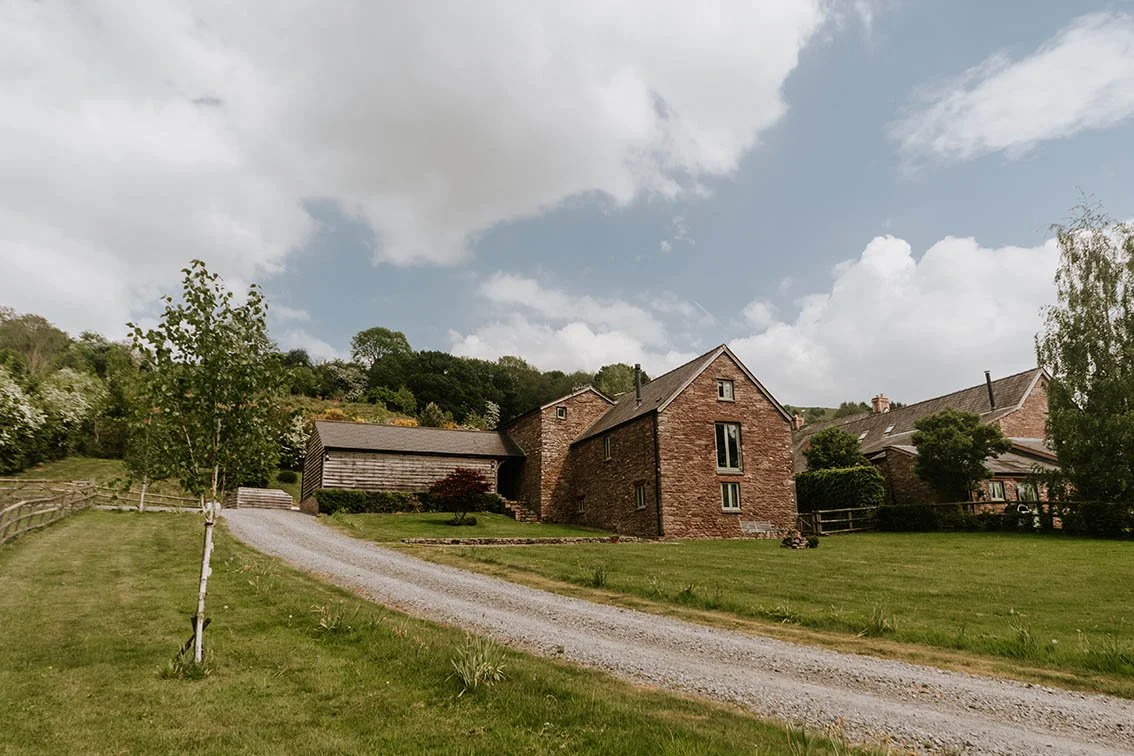 A rustic house with a driveway leading up to it, surrounded by green grass, trees, and a partly cloudy sky.