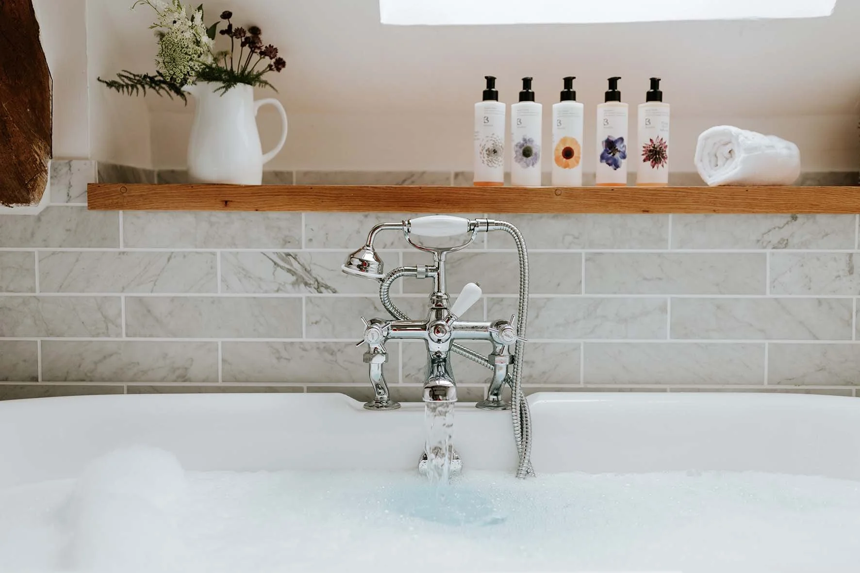 A bathtub with bubbly water and a vintage-style silver faucet, set against a tiled wall. On a wooden shelf above, there is a white pitcher with flowers, bottles of bath products, a rolled white towel, and a small window.