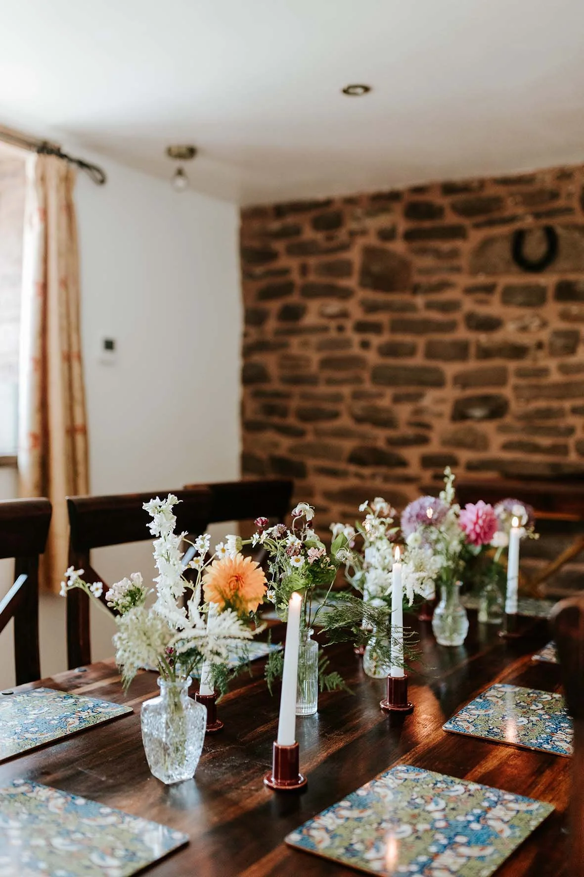 A dining table decorated with vases of flowers and lit candles in a cozy room with a brick wall and a window with curtains.