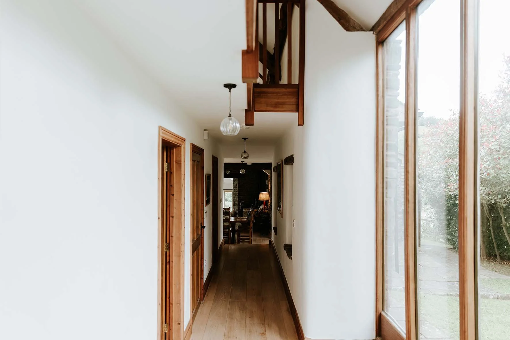 Interior view of a hallway with wooden trim, doors on the left, large windows on the right, and a staircase overhead. At the end of the hall, a dining table and chairs with a lamp are visible.