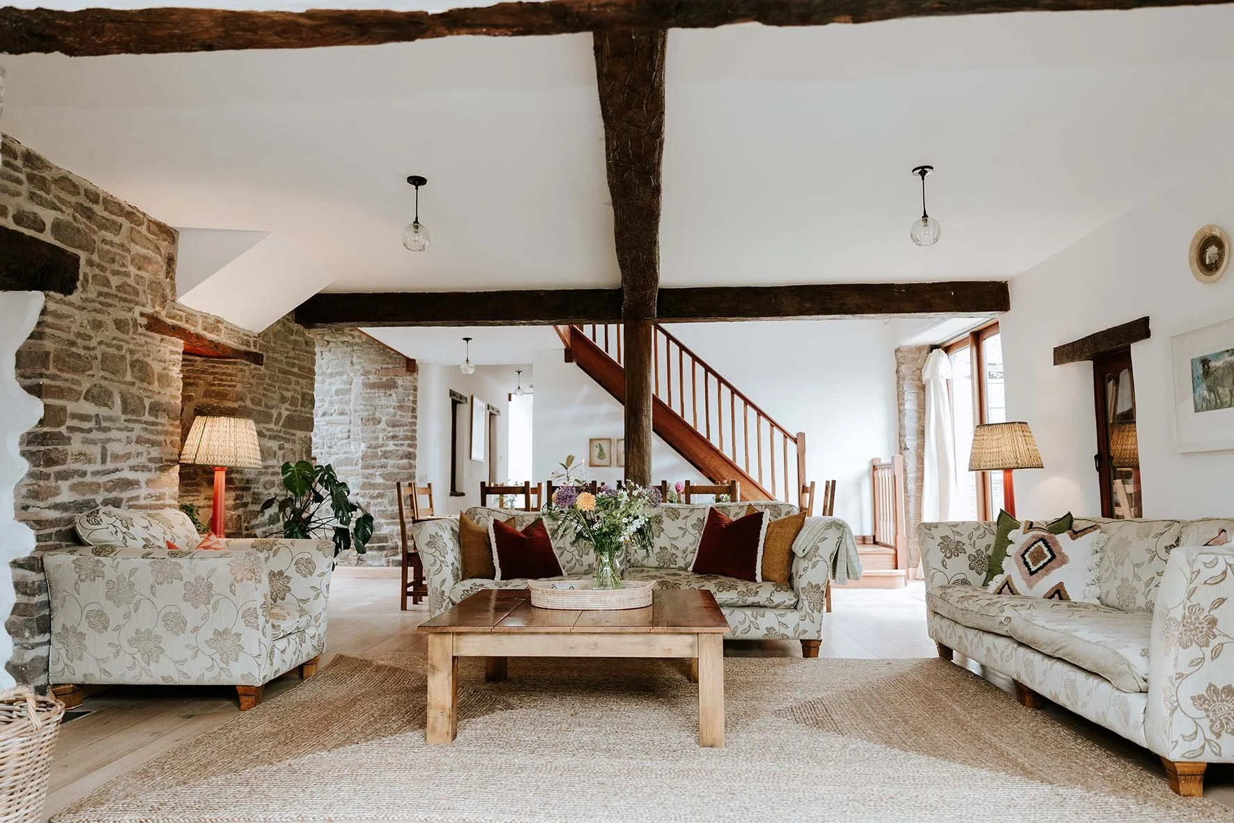 Living room with stone walls, wooden beams, floral-patterned sofas, a wooden coffee table with a flower arrangement, dining area in the background, and natural light from windows.