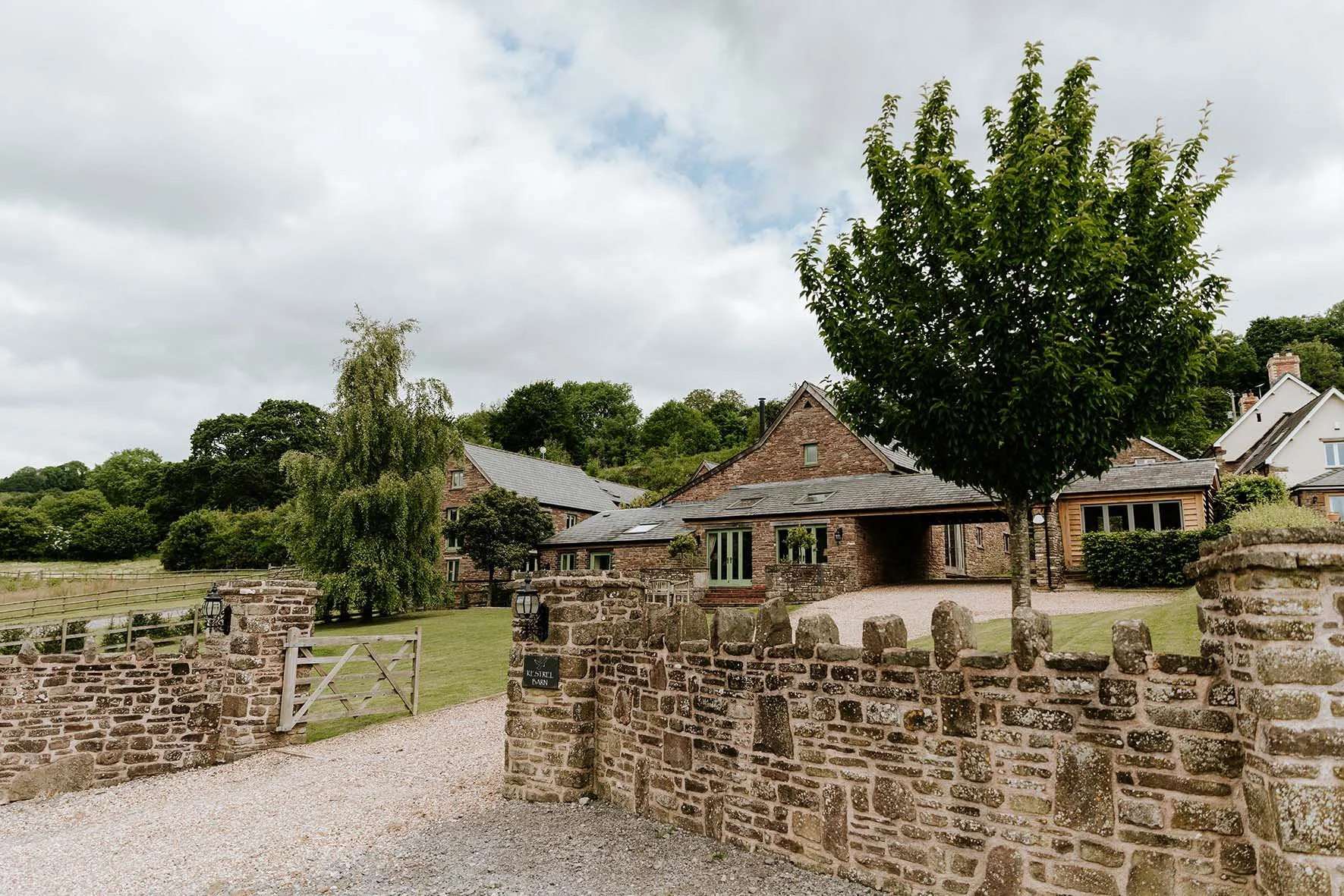 View of a rustic brick house with a driveway, surrounded by trees and a stone fence with lanterns on top, under a partly cloudy sky.
