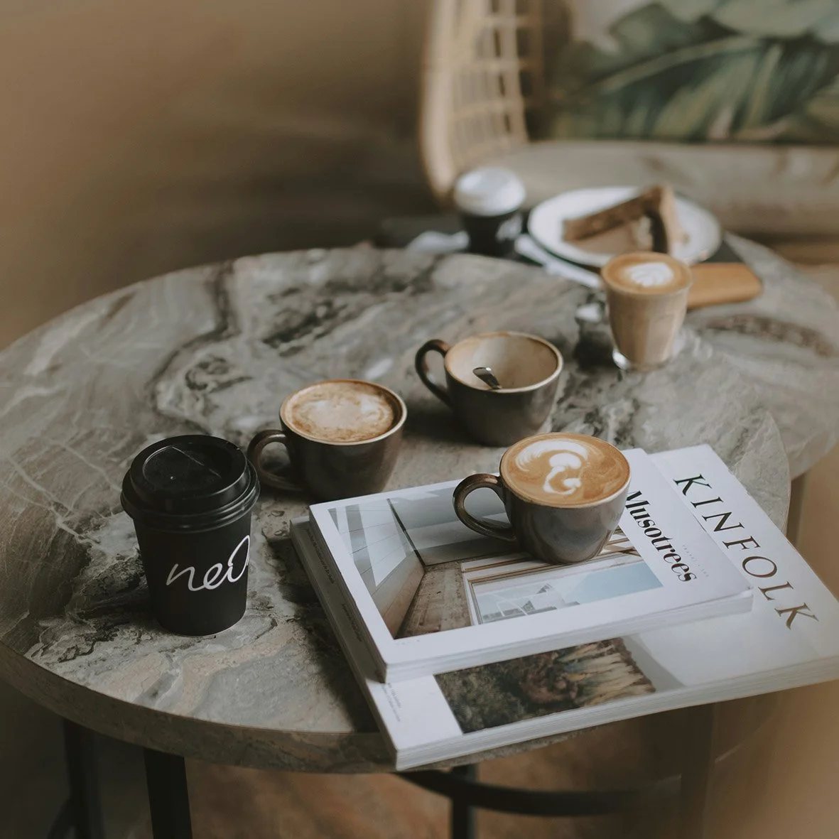A marble-top round table with four cups of coffee, a black to-go coffee cup, and magazines, with a small plate and snacks in the background.