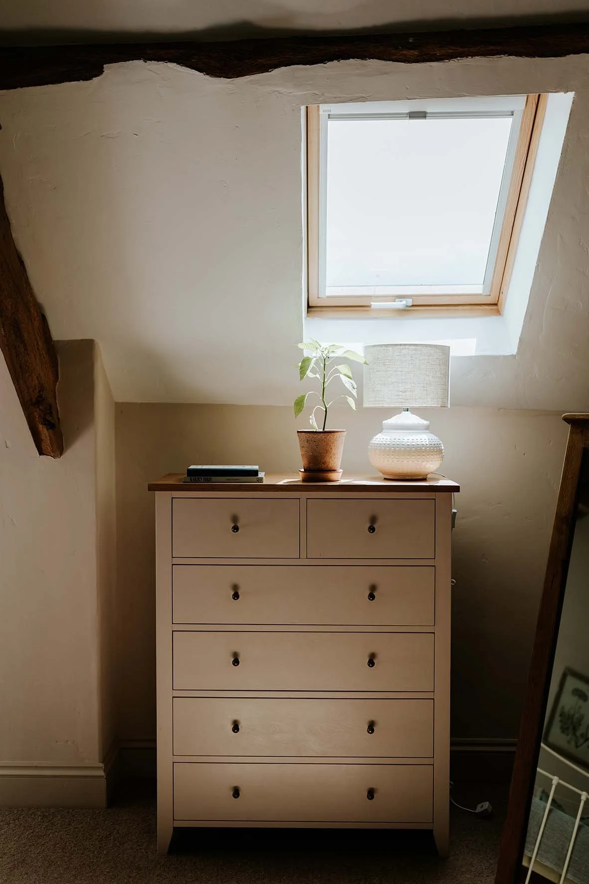 A beige dresser with five drawers is placed beneath a skylight window, with a potted plant, a white textured lamp, and a closed book on top.