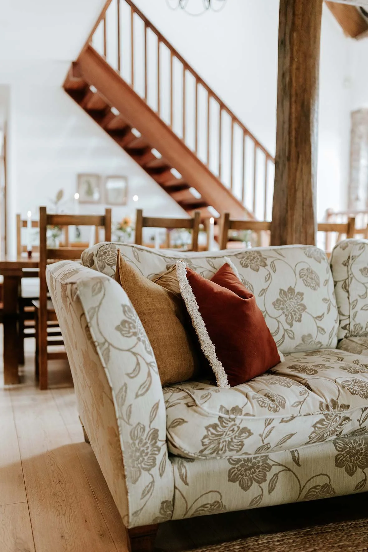 Living room with a floral patterned beige sofa, decorative pillows in brown and rust, a wooden dining table with chairs, a wooden staircase, and a chandelier in the background.