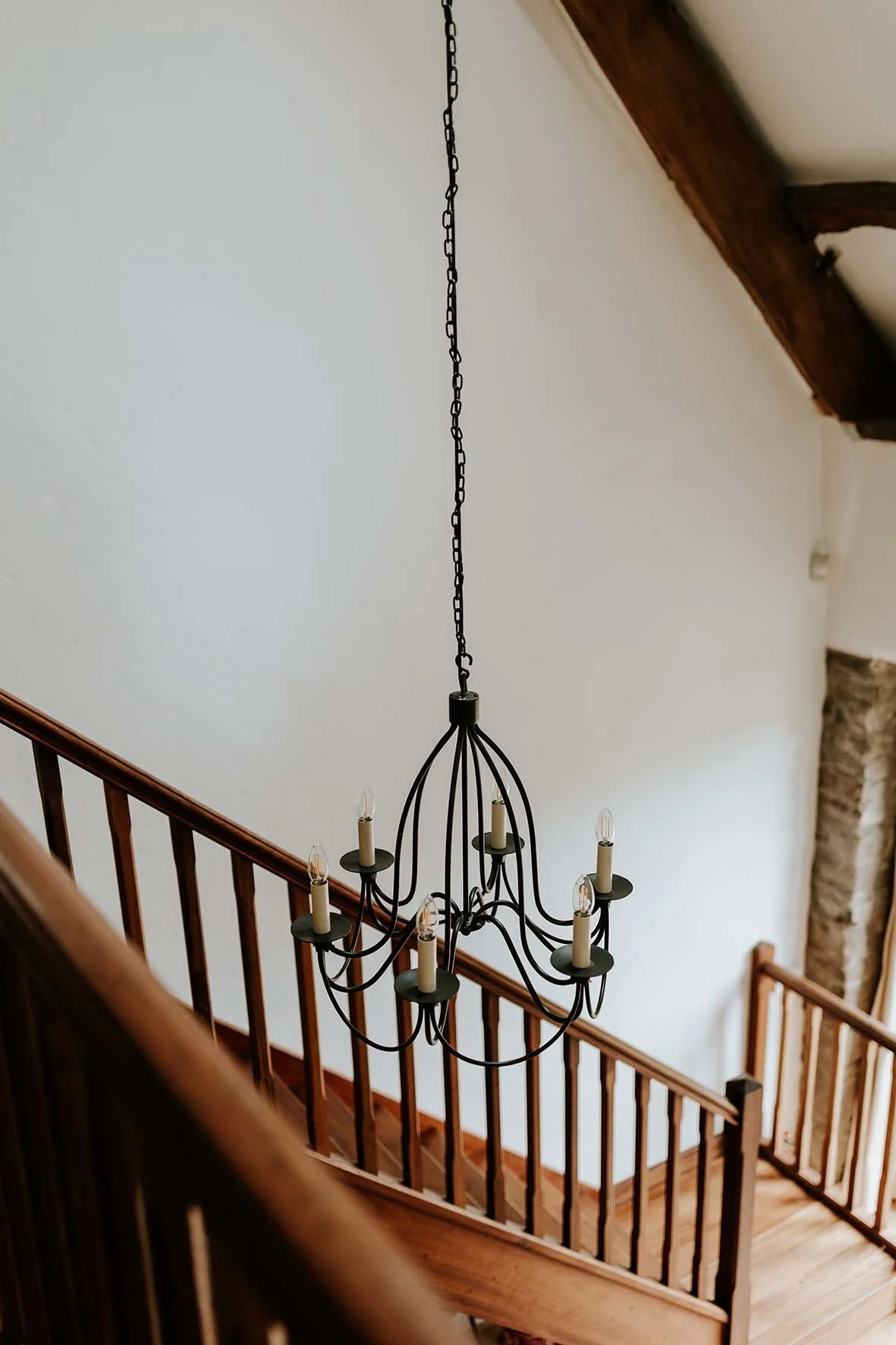 A black chandelier with eight light bulbs hanging above a wooden staircase with a wooden handrail in a house interior.