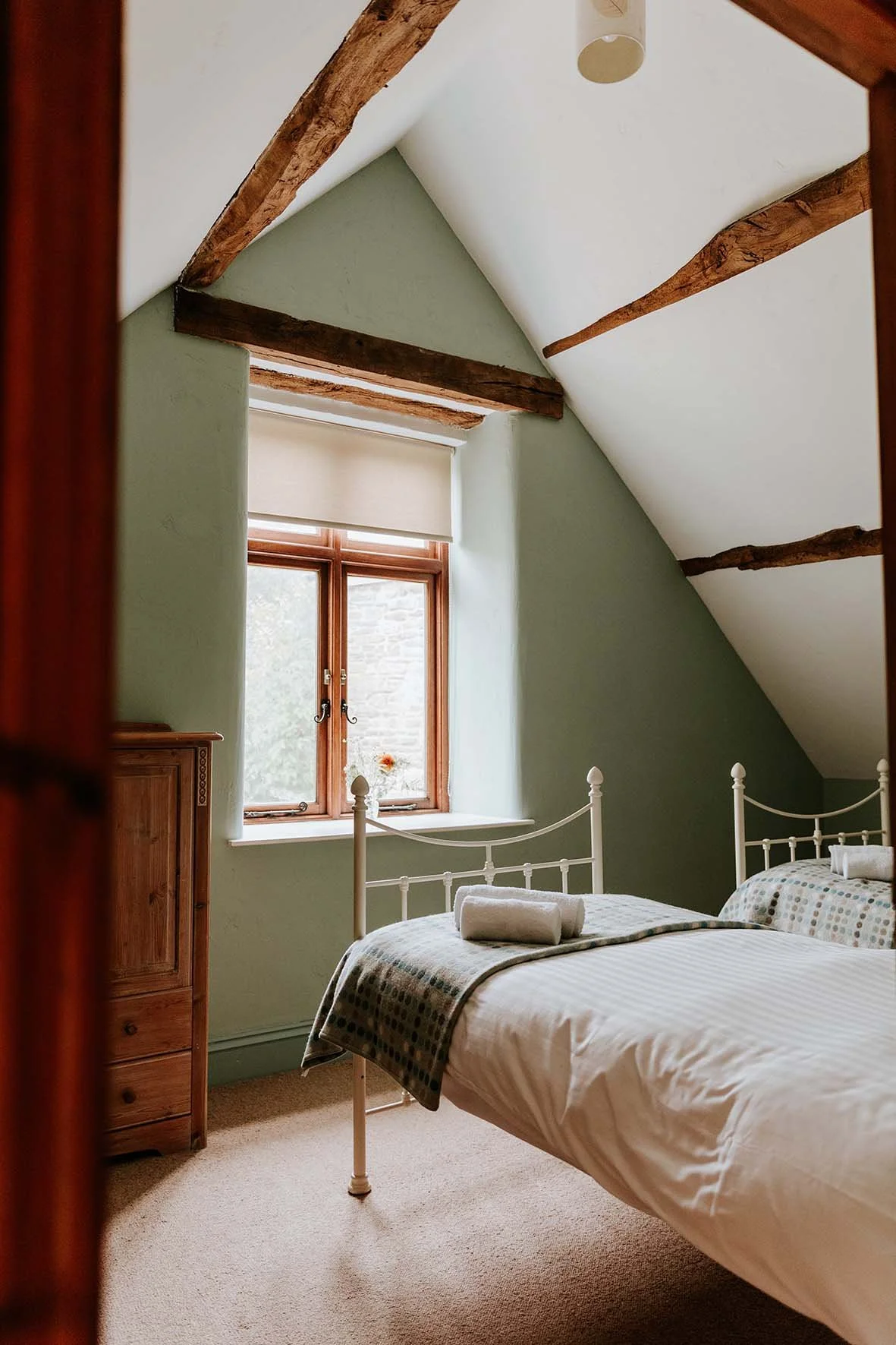 A cozy bedroom with a white metal bed frame, a neatly made bed with a folded towel on top, and a window with natural light coming in. The room has soft green walls, wooden beams on the ceiling, and a beige carpet.