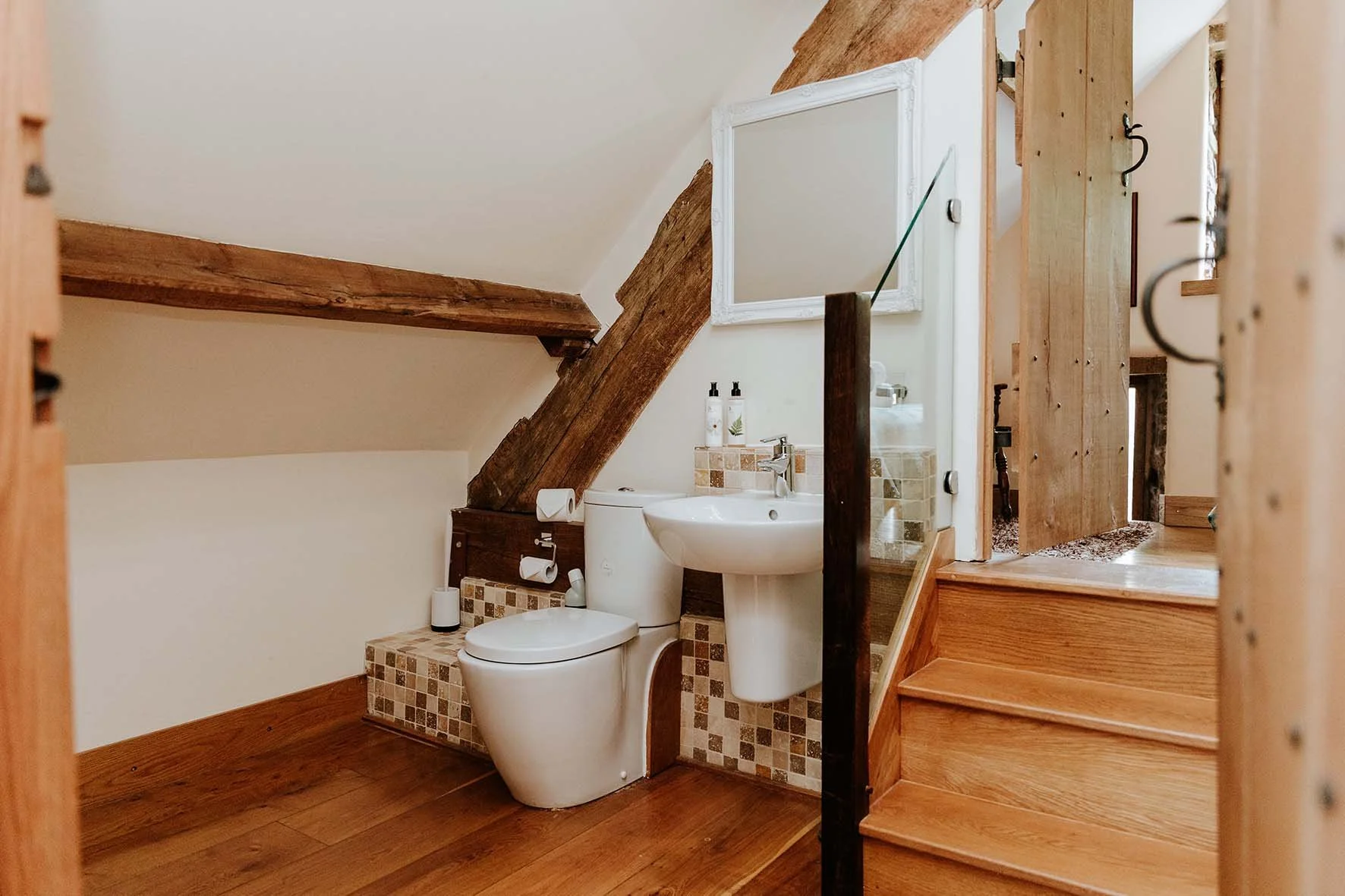 Small bathroom with a modern toilet, a white sink, and a mirror, under a sloped ceiling with wooden beams, featuring wooden stairs and flooring.