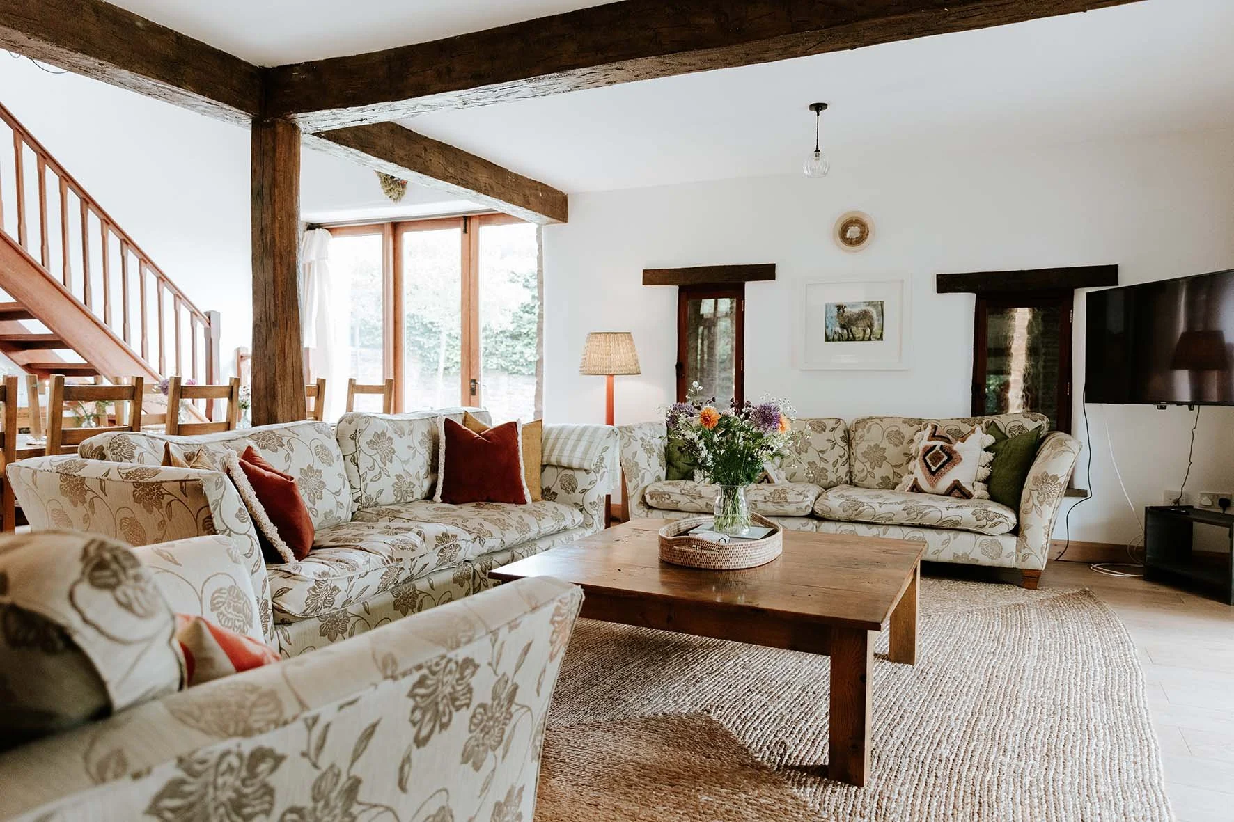 Living room with floral sofas, a wooden coffee table with a vase of flowers, a fireplace, a television, and large windows letting in natural light. Wooden beams are visible on the ceiling.