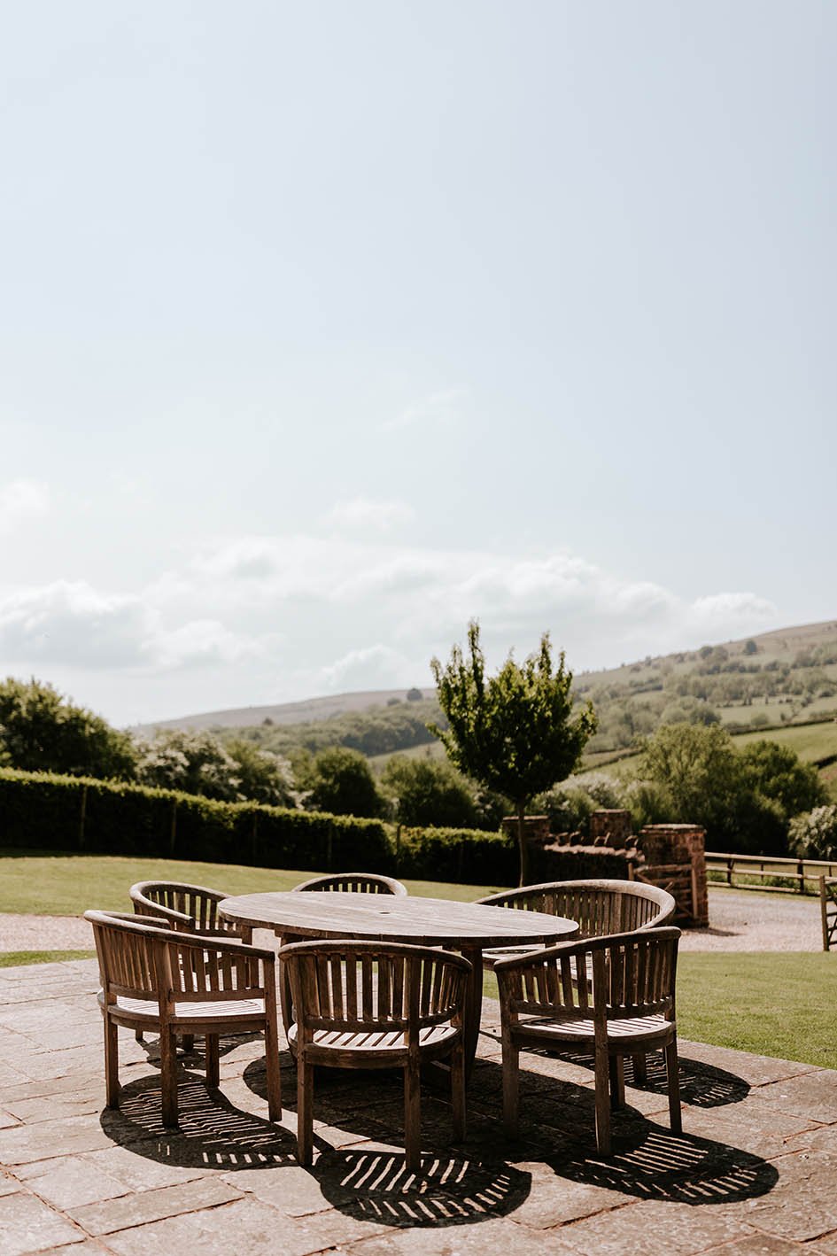 Circular wooden outdoor table with six matching chairs on a stone patio, overlooking a green landscape with trees and rolling hills under a partly cloudy sky.