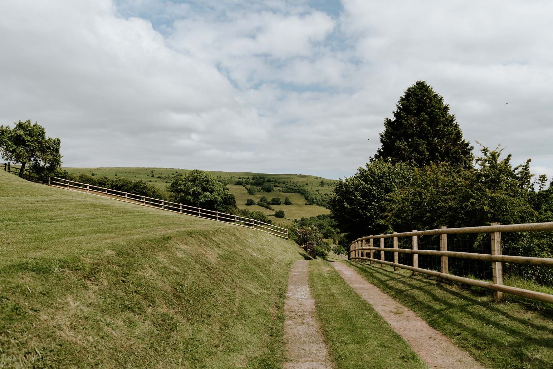 A dirt pathway runs through rolling green hills, bordered by wooden fences on both sides, with scattered trees and a cloudy sky overhead.