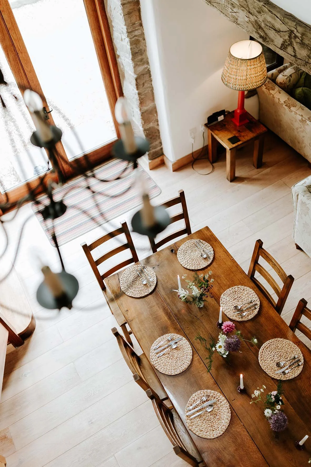 A high-angle view of a wooden dining table set for dinner with six woven placemats, silverware, and small candles, in a cozy room with light wood flooring, a floral centerpiece, a side table with a lamp, and a large glass door with curtains.