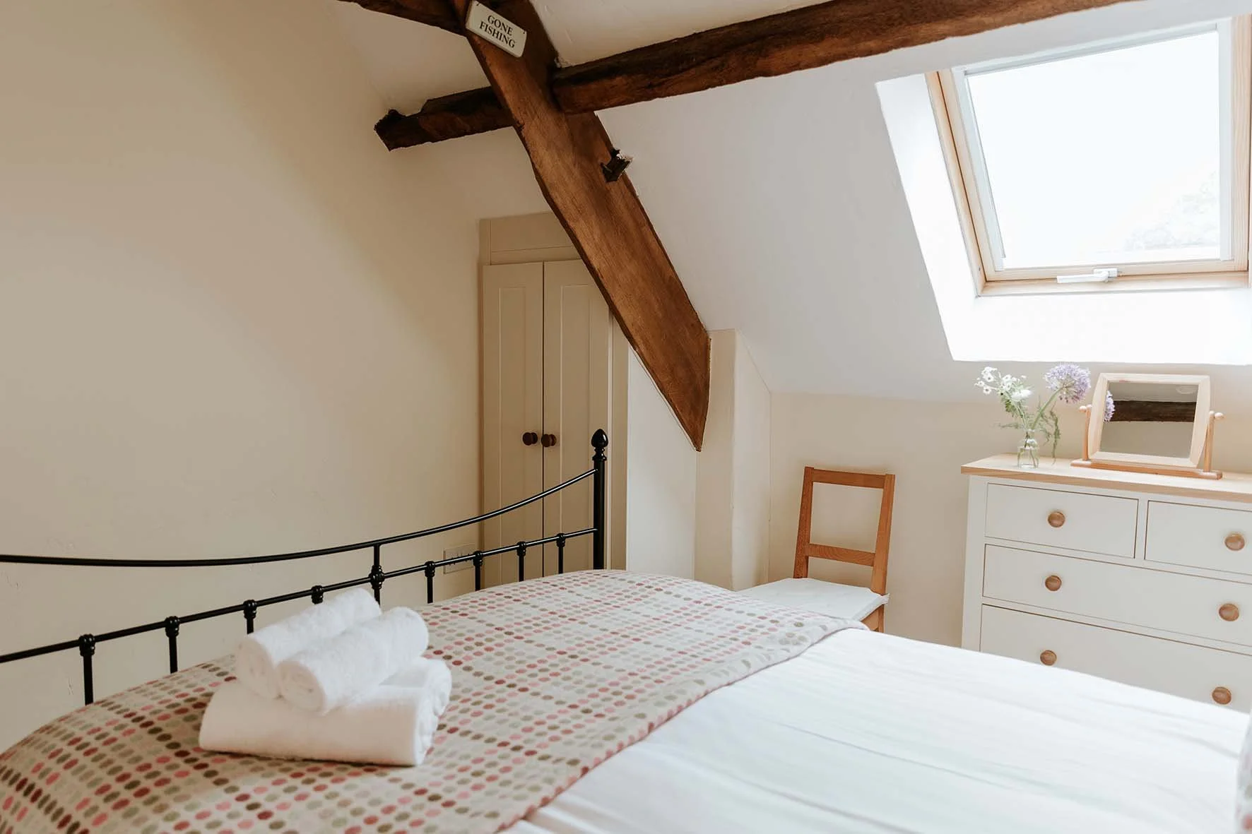 A cozy bedroom with a metal bed frame, folded towels on the bed, a white dresser with a mirror, a chair, and a skylight window in the ceiling.