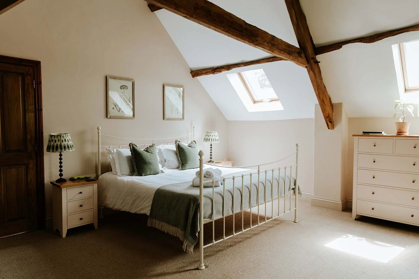 A cozy bedroom with a white metal bed frame, green pillows, a green throw blanket, and rolled towels on the bed. Two bedside tables with patterned lamps, a wooden dresser with a potted plant, and two skylight windows in a room with sloped ceilings an