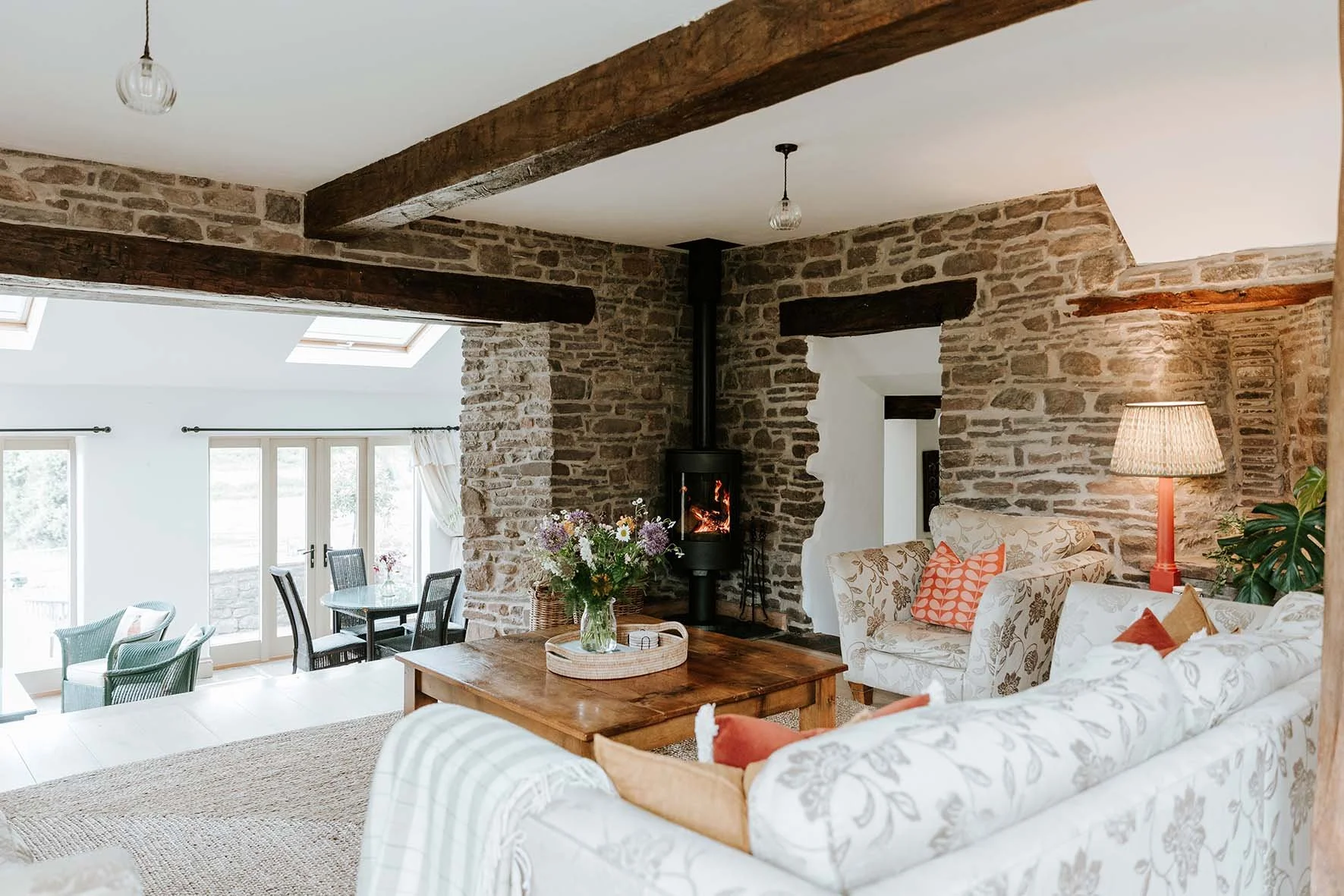 Living room with brick walls and ceiling beams, a wood stove with fire, floral-patterned sofas, a wooden coffee table with a flower vase, a lamp with a shade, and a dining area with sliding glass doors leading outside.