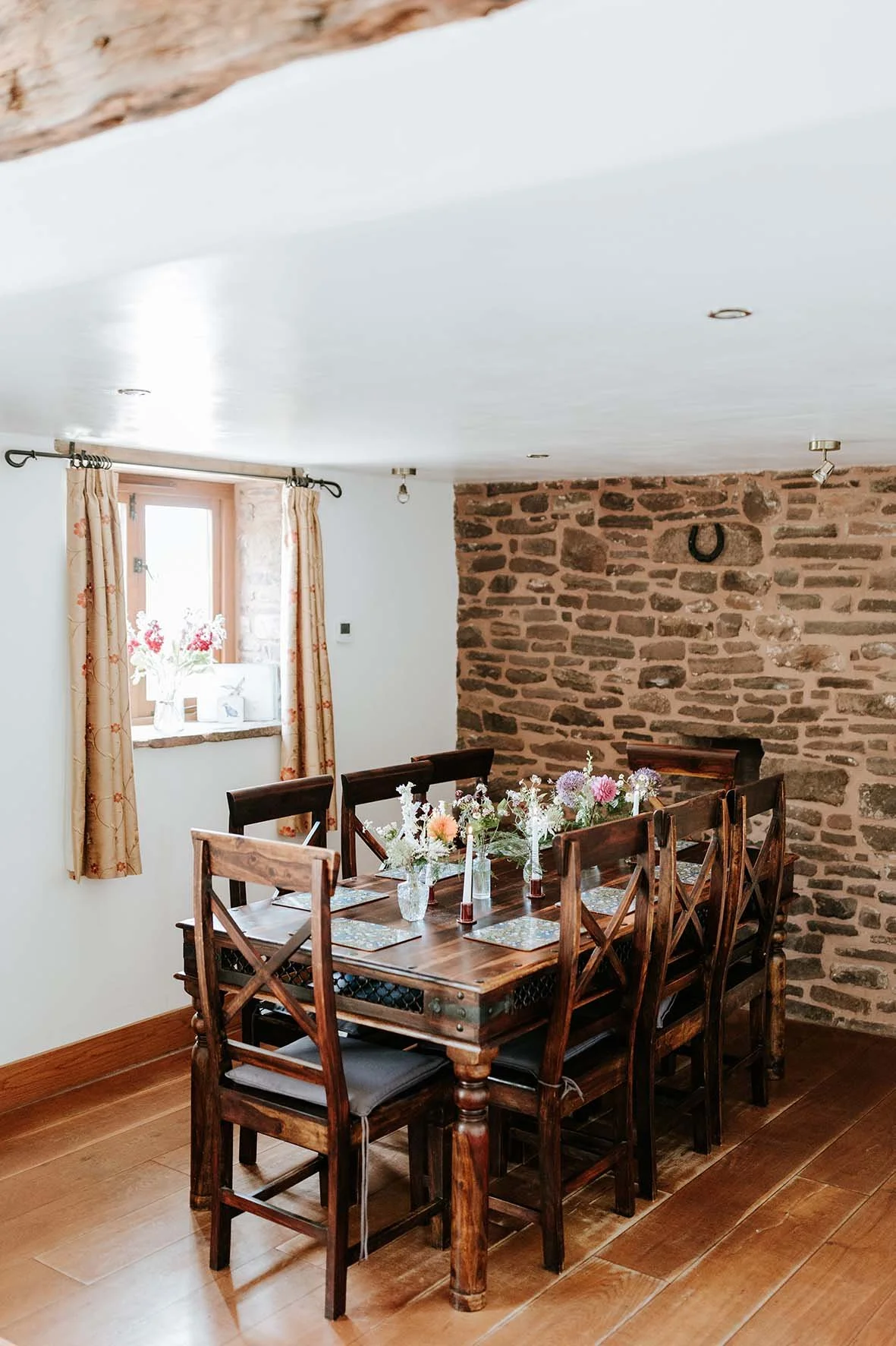 A cozy dining room with a rustic brick wall, wooden table with floral arrangements, six wooden chairs, a window with cream-colored floral curtains, and a white ceiling with track lighting.