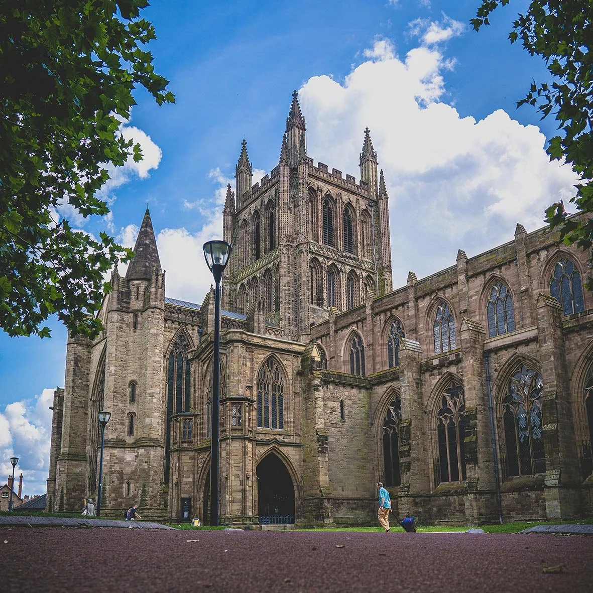 A large, historic cathedral with gothic architecture, tall spires, and tall arched windows, seen from a low angle with a blue sky and clouds above, surrounded by trees and a few people walking nearby.