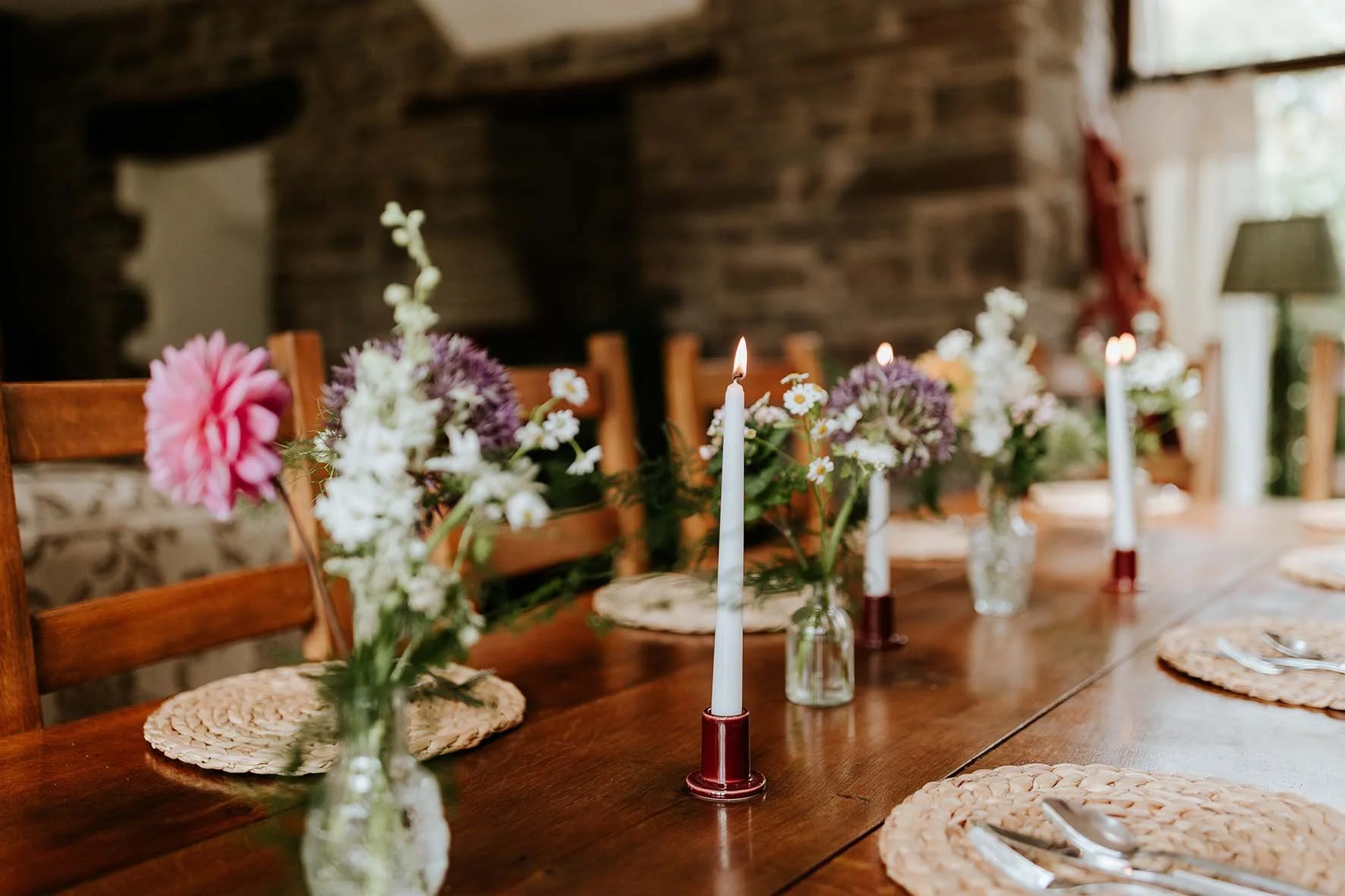 A wooden dining table decorated with pink and white flowers in small glass vases, lit white candles in candleholders, woven placemats, and silverware, in a cozy room with a brick wall and window in the background.