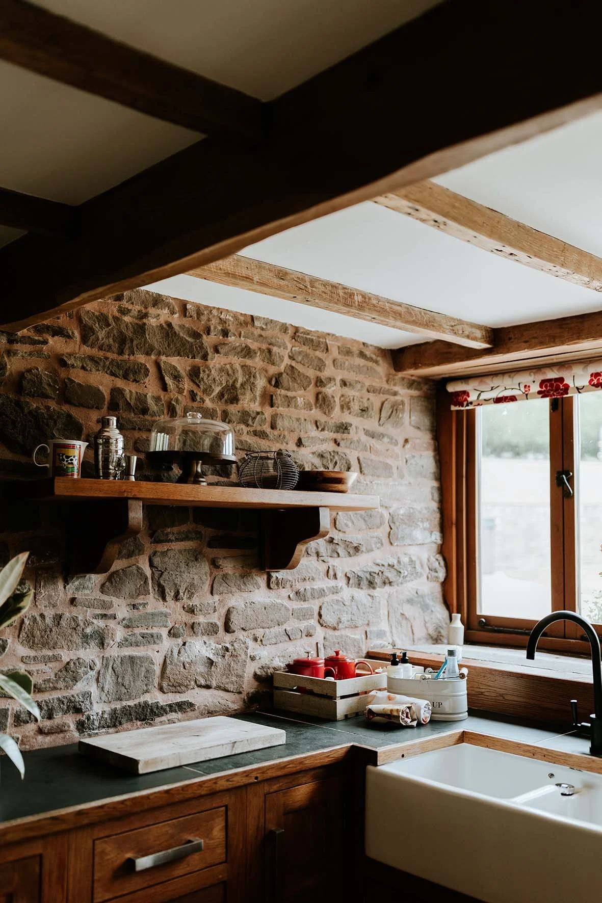A rustic kitchen with a stone wall, wooden cabinets, and a window with a floral valance. The countertop has red and white kitchenware, a cutting board, a soap dispenser, and a sink with a black faucet.