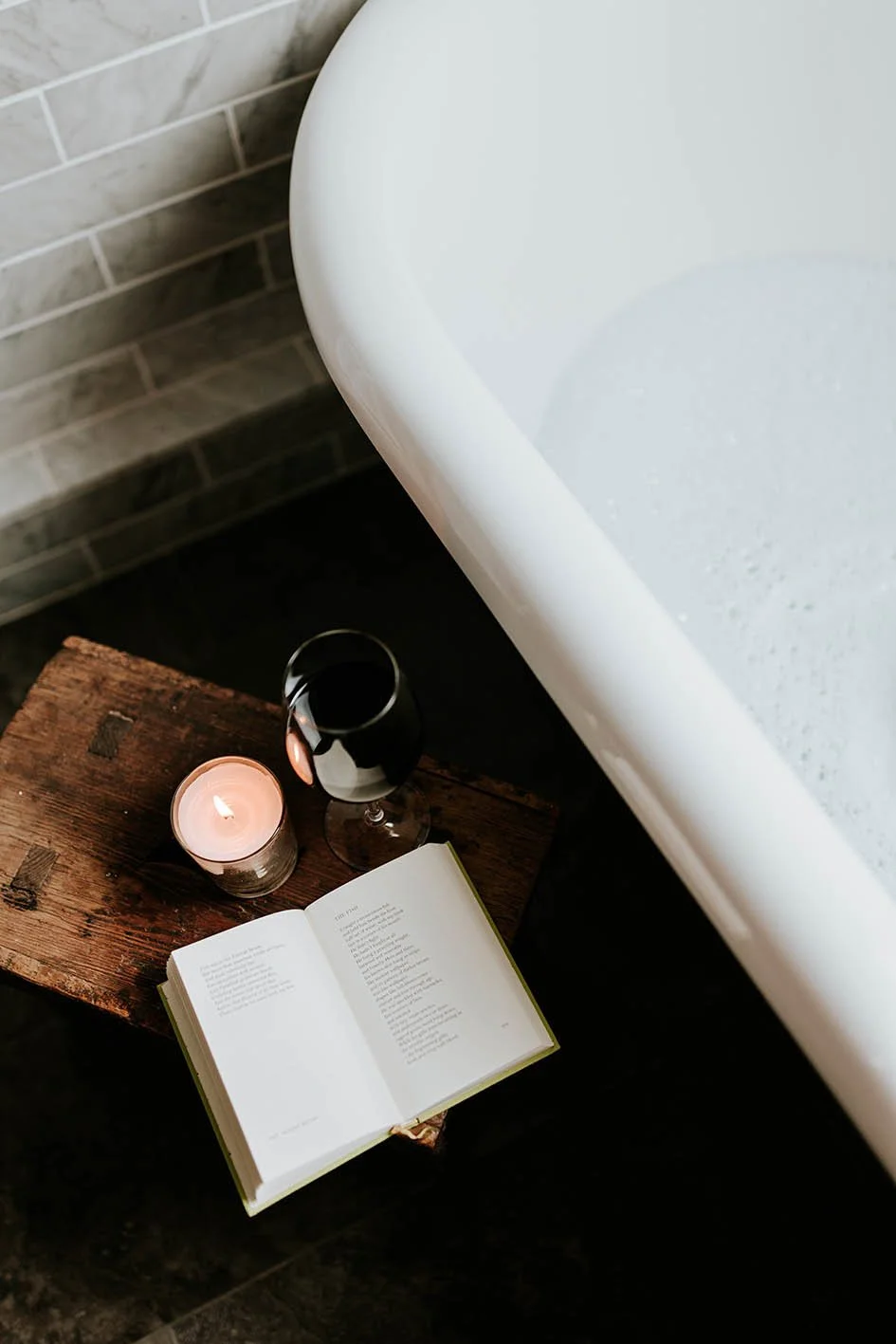 A cozy bathroom scene with a bathtub, an open book, a lit candle, and a glass of red wine on a rustic wooden stool.