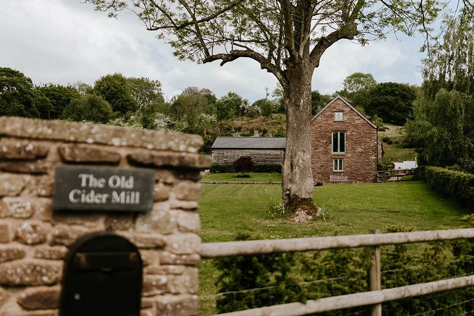A rural scene with a stone sign that reads 'The Old Cider Mill,' a wooden fence, and a large tree in the foreground. In the background, there is a brick house, a wooden shed, and green trees under a cloudy sky.